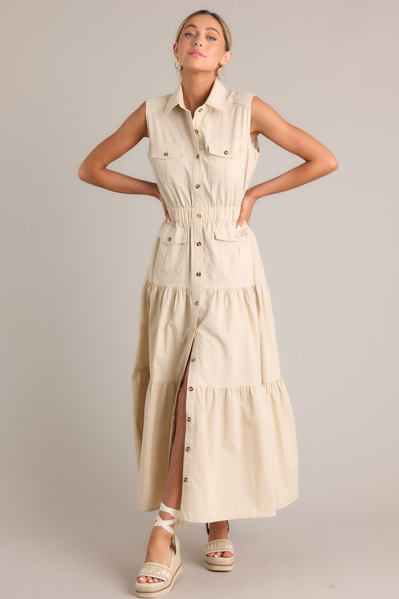 This beige maxi dress features a collared neckline, a full button front, buttoned breast pockets, a thick elastic waistband, buttoned hip pockets, and a tiered design.