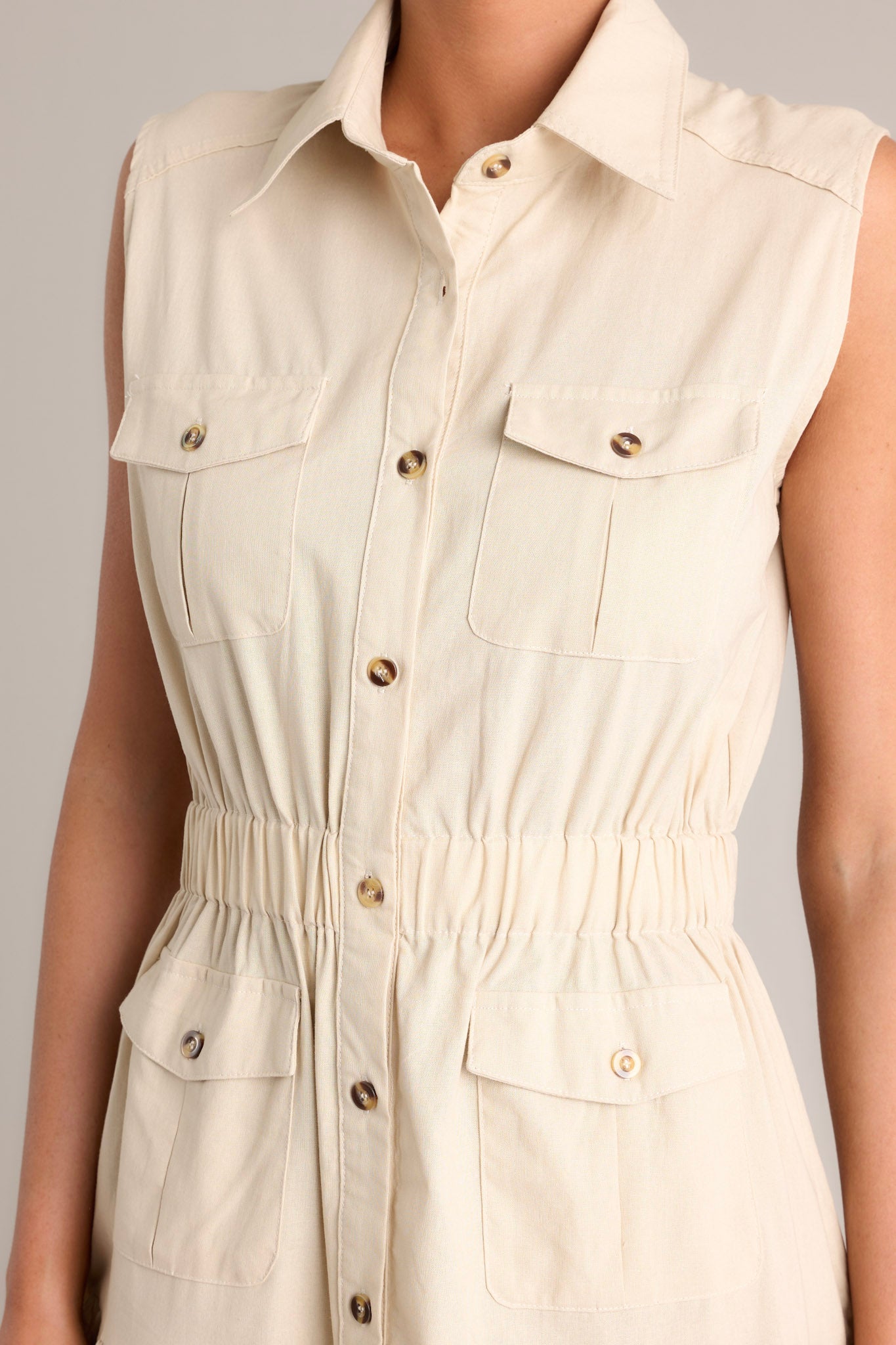 Close up view of this beige maxi dress that features a collared neckline, a full button front, buttoned breast pockets, a thick elastic waistband, buttoned hip pockets, and a tiered design.