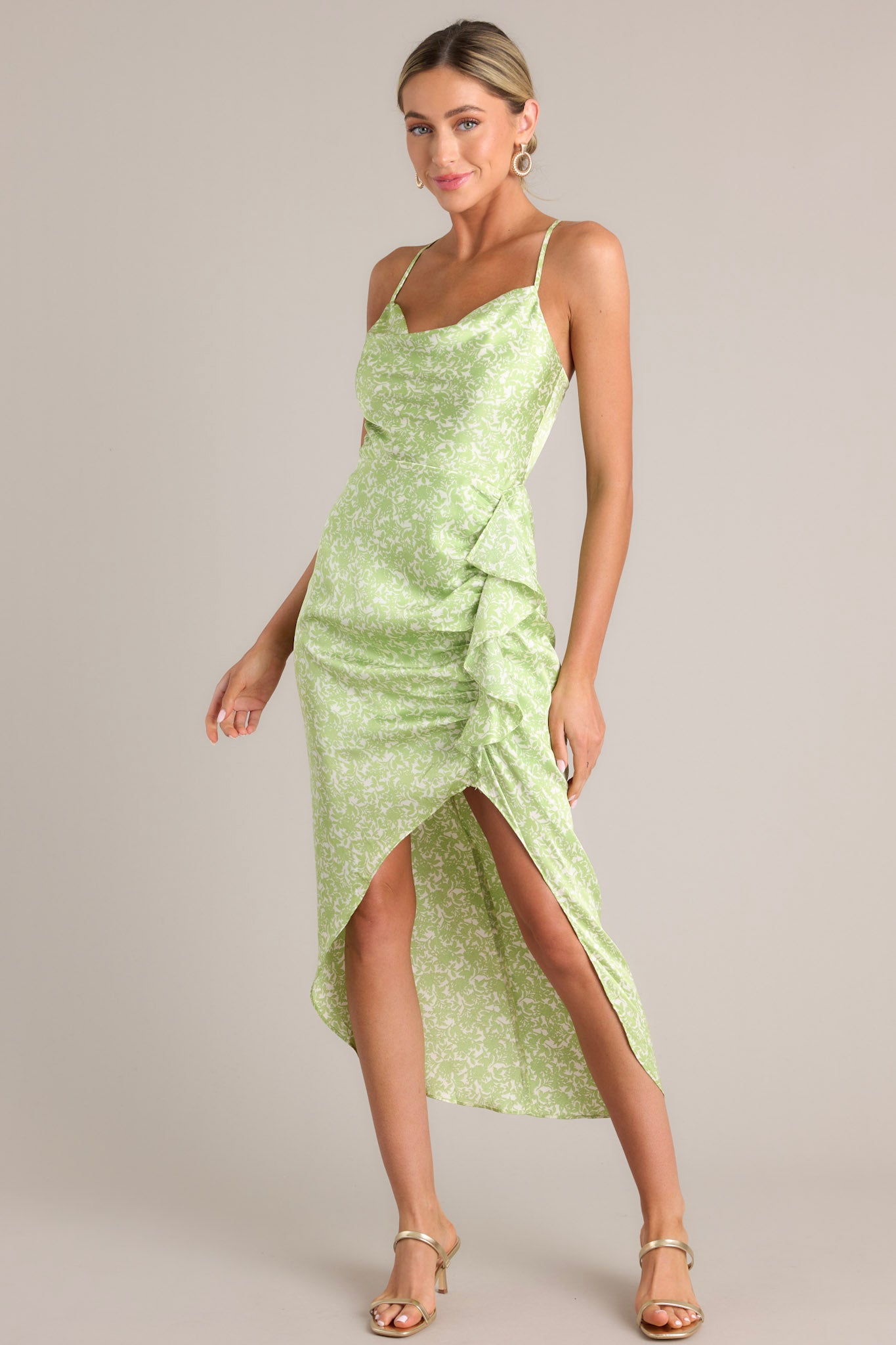 Angled full body view of this dress that features a cowl neckline, thin adjustable crossed straps, a zipper down the back, a ruffled hip feature, and an asymmetrical hemline.