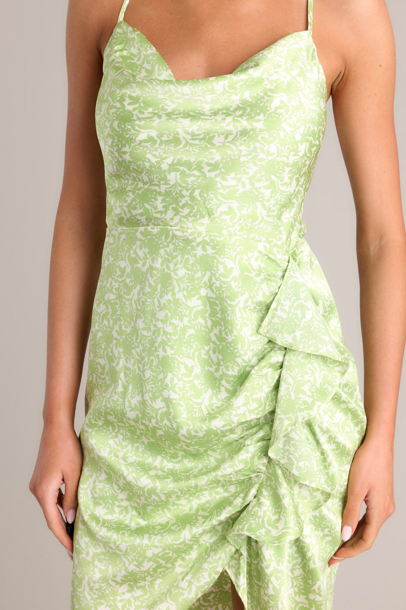 Close up view of this dress that features a cowl neckline, thin adjustable crossed straps, a zipper down the back, a ruffled hip feature, and an asymmetrical hemline.