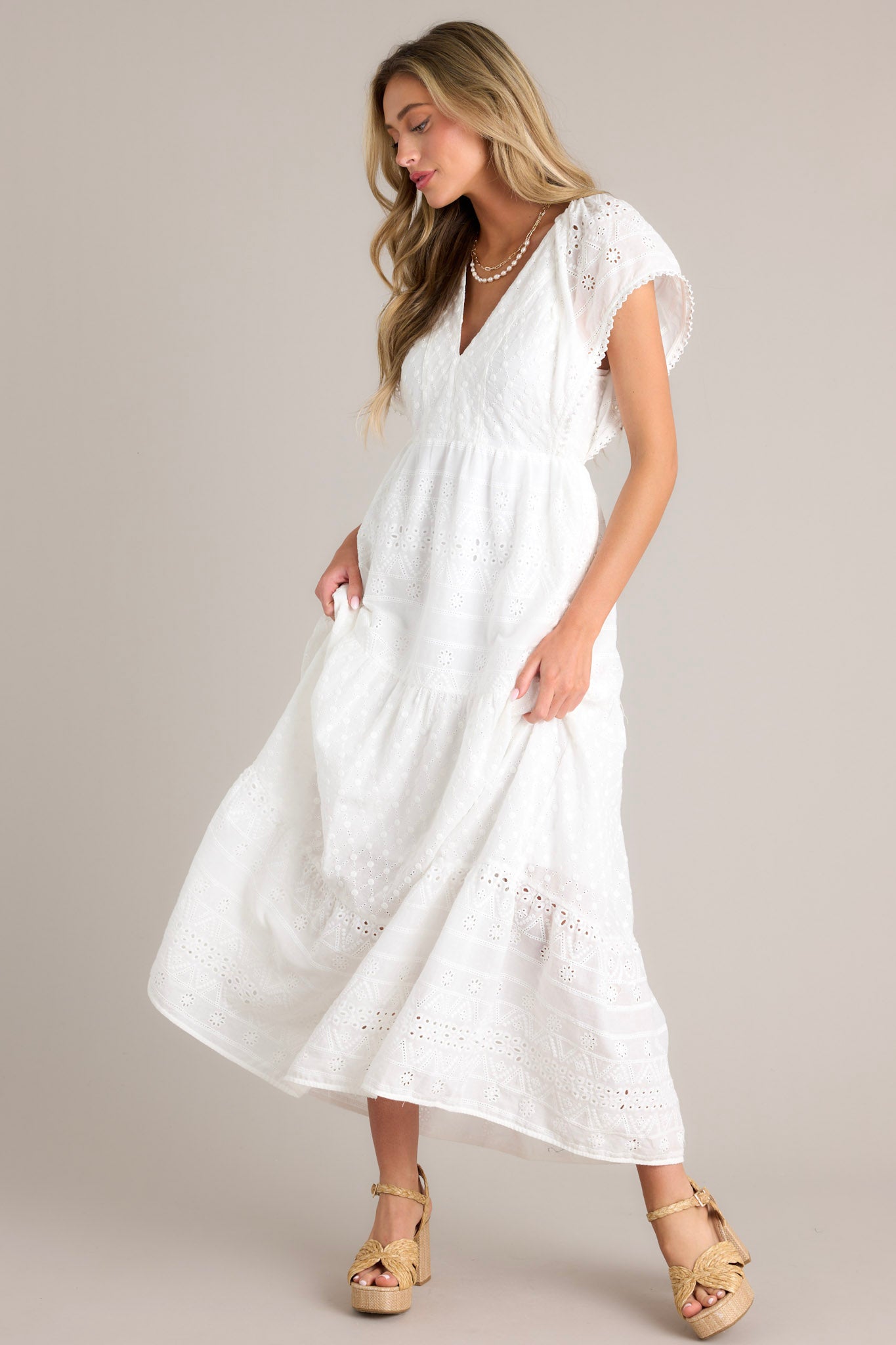 Angled view of this white maxi dress with v-neckline, removable slip, thin elastic waistband, hip pockets, eyelet detailing, and faux pearl trimmed sleeves.