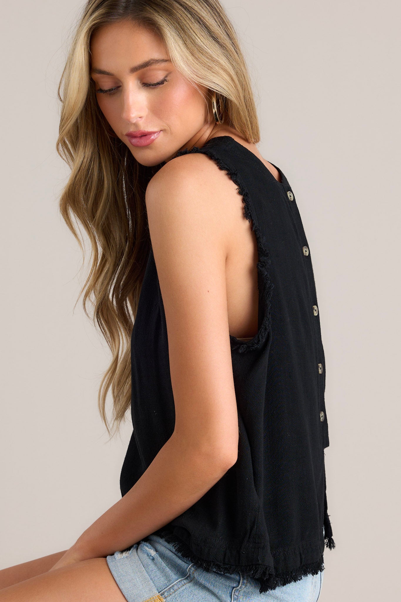 Side view of this black tank that features a crew neckline, functional buttons down the back, frayed shoulders, and a frayed hemline.