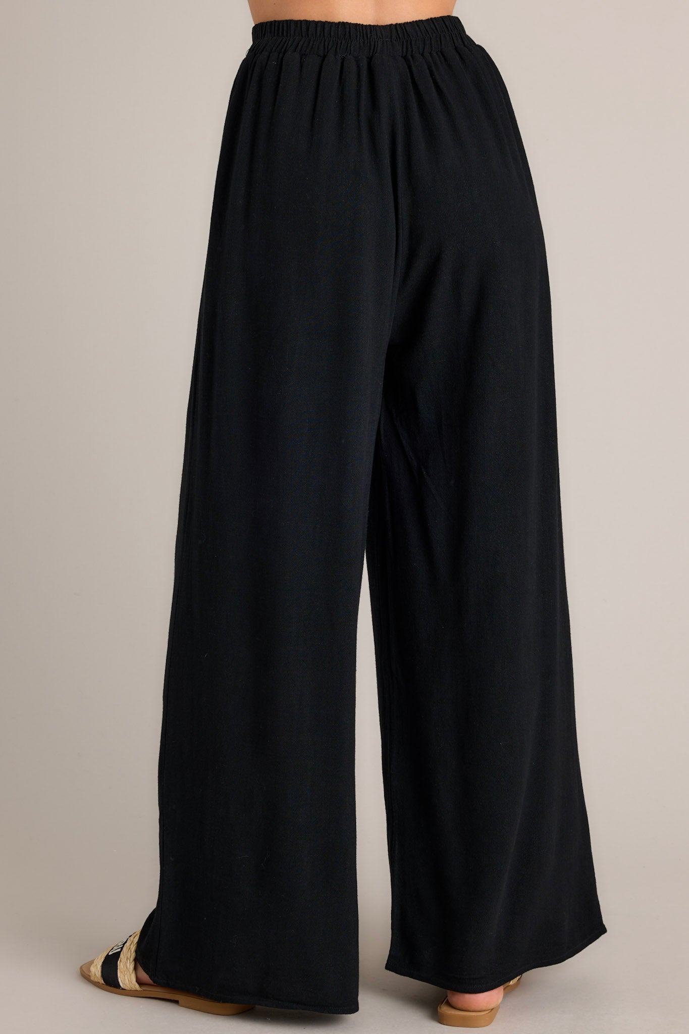 Back view of these black pants that feature a high waisted design, an elastic waistband, a self-tie waist feature, and a wide leg.