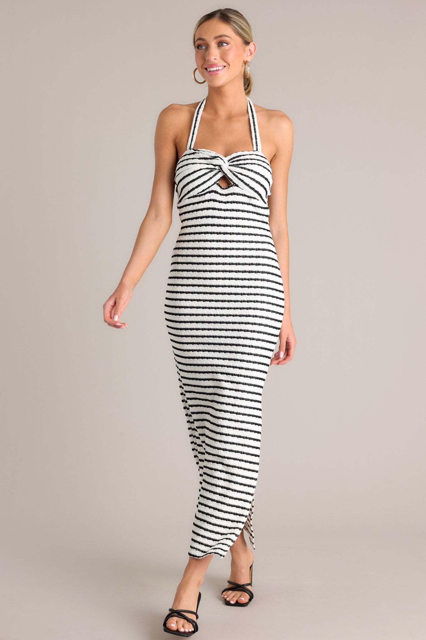 Angled full body view of this stripe maxi dress that features a self-tie halter neckline, twist detailing in the bust, a chest cutout, an open back, a textured material, and a side slit.