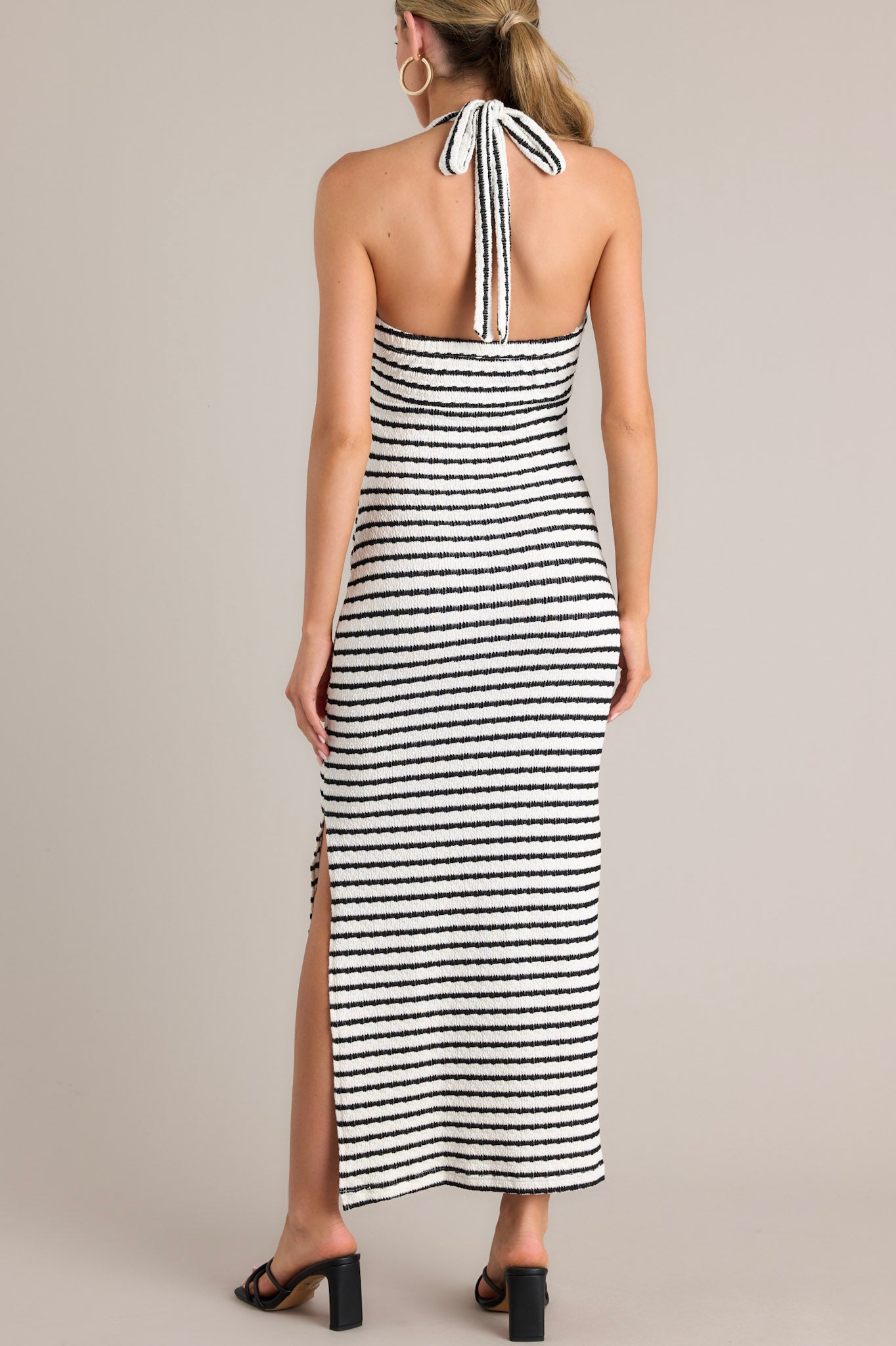 Back view of this stripe maxi dress that features a self-tie halter neckline, twist detailing in the bust, a chest cutout, an open back, a textured material, and a side slit.