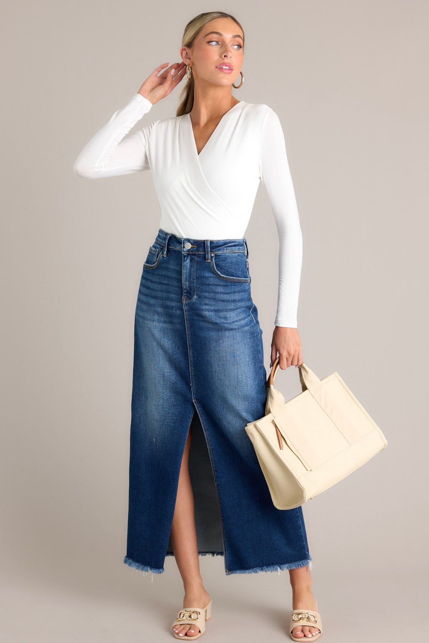 Angled full body view of this dark wash denim midi skirt featuring a high waisted design, belt loops, a zipper and button closure, functional pockets, a 15" slit up the center, and raw hem detailing.