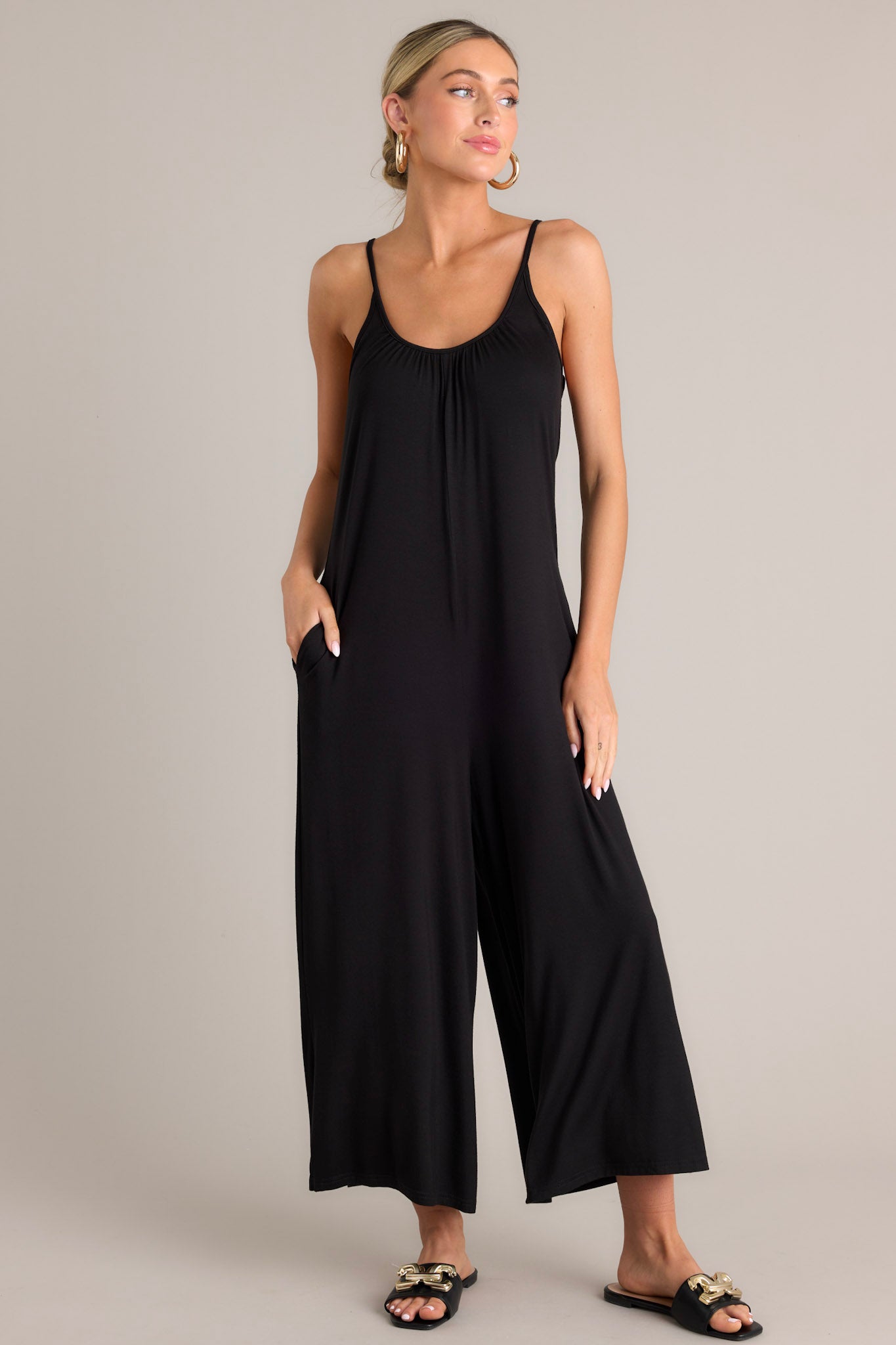 Angled full body view of this black jumpsuit that features a scoop neckline, thin adjustable straps, an open back, a relaxed fit, functional hip pockets, and a wide leg.