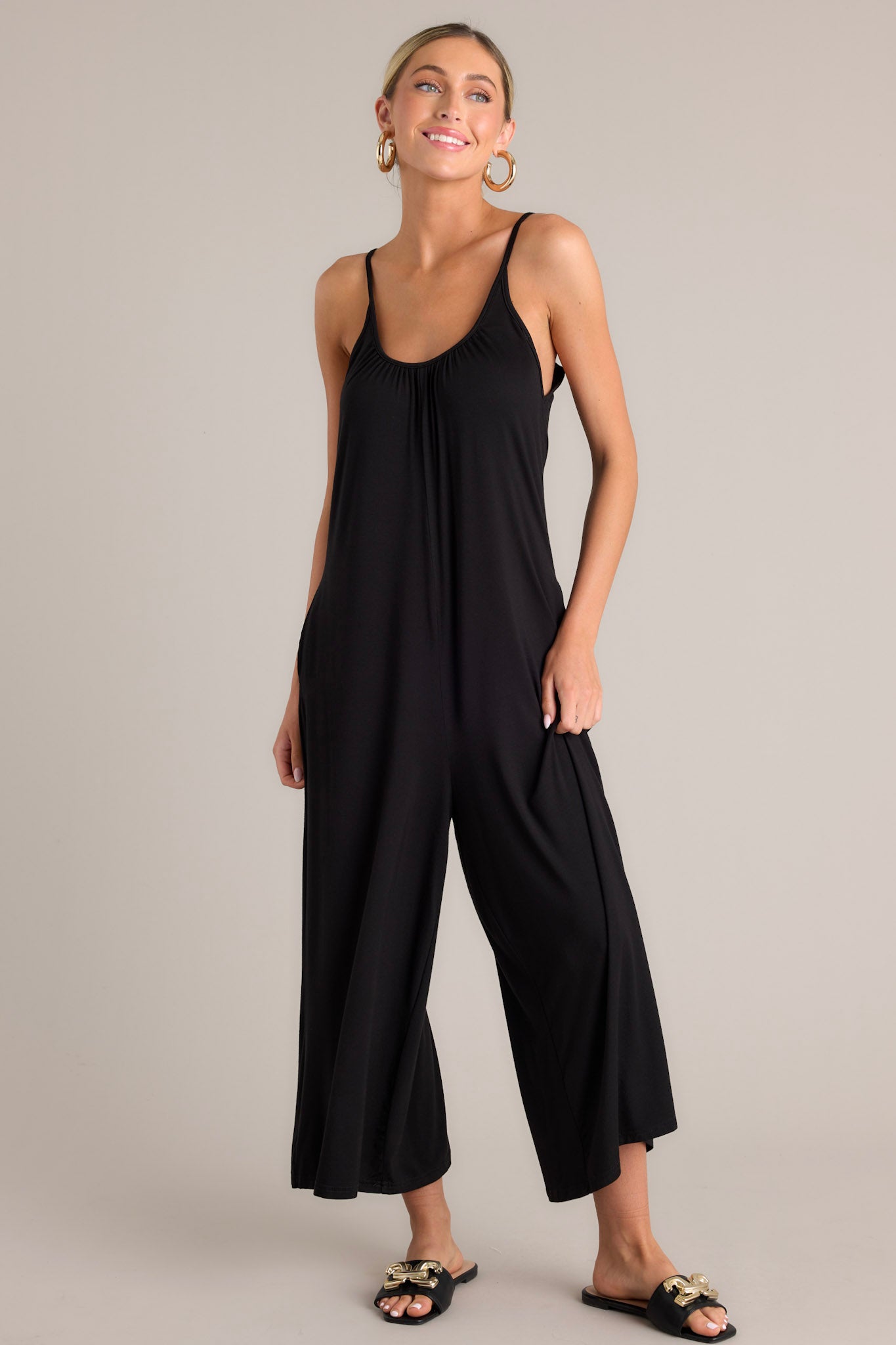 Front view of this black jumpsuit that features a scoop neckline, thin adjustable straps, an open back, a relaxed fit, functional hip pockets, and a wide leg.