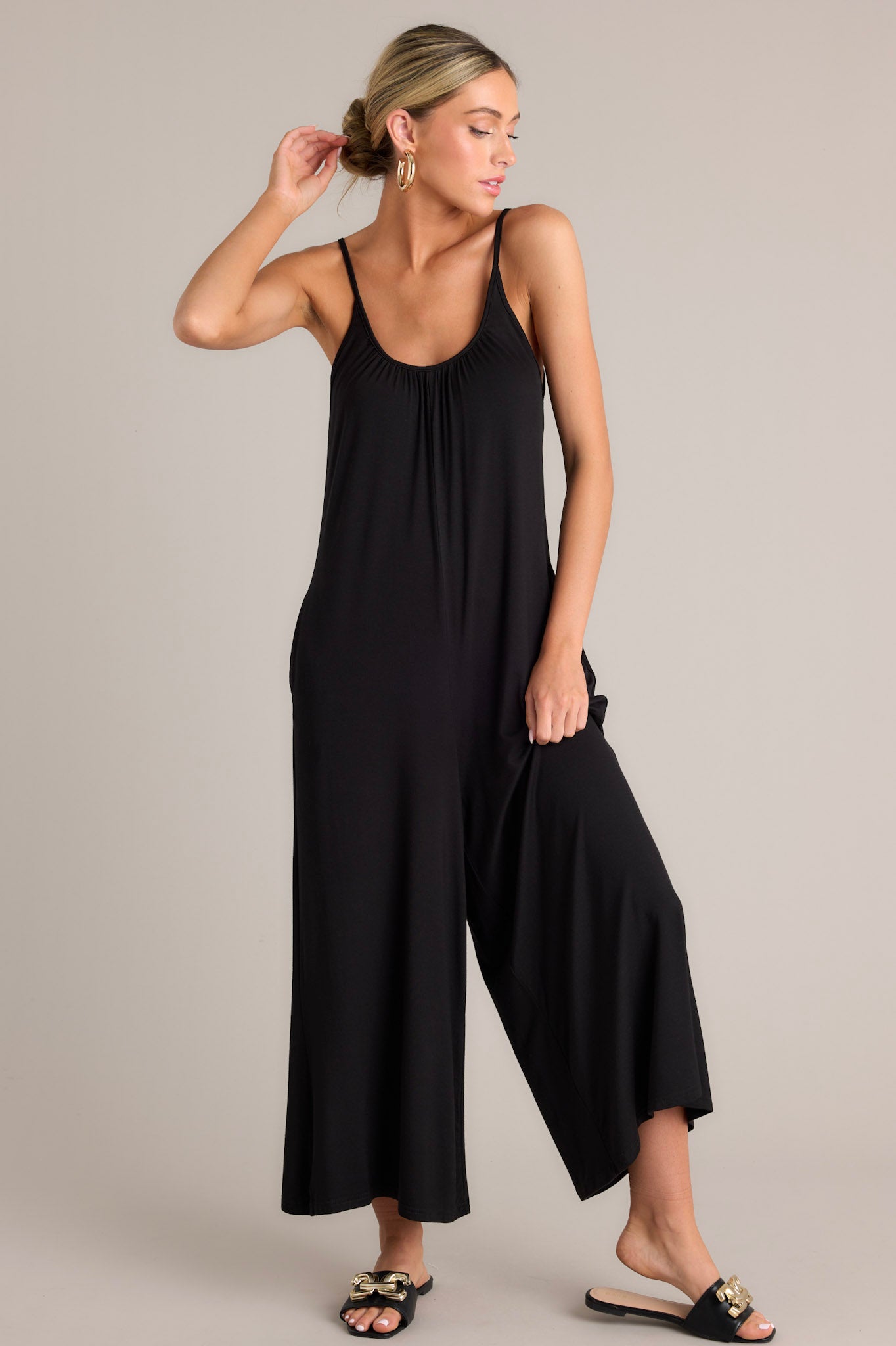 Angled full body view of this black jumpsuit that features a scoop neckline, thin adjustable straps, an open back, a relaxed fit, functional hip pockets, and a wide leg.