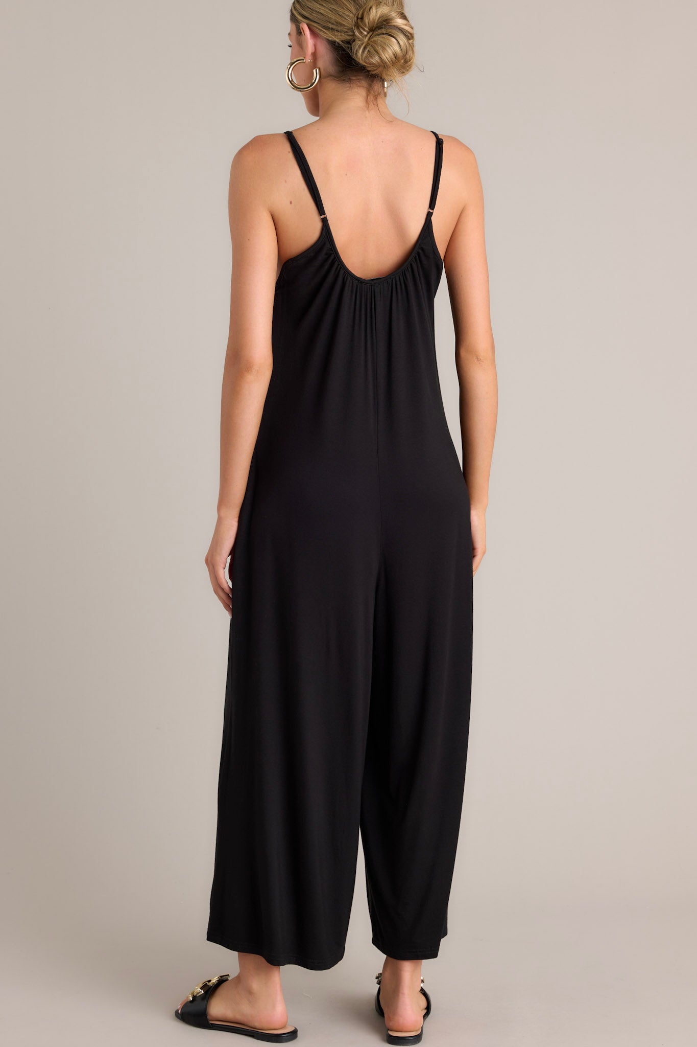 Back view of this black jumpsuit that features a scoop neckline, thin adjustable straps, an open back, a relaxed fit, functional hip pockets, and a wide leg.