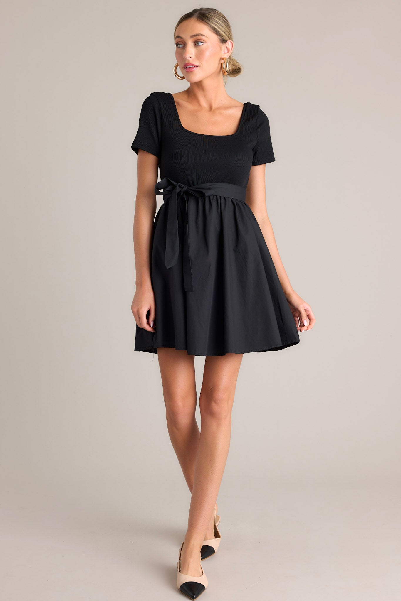 Angled full body view of this black mini dress that features a square neckline, a ribbed bodice, an elastic waistband, belt loops, a self-tie waist belt, and a flowing silhouette.