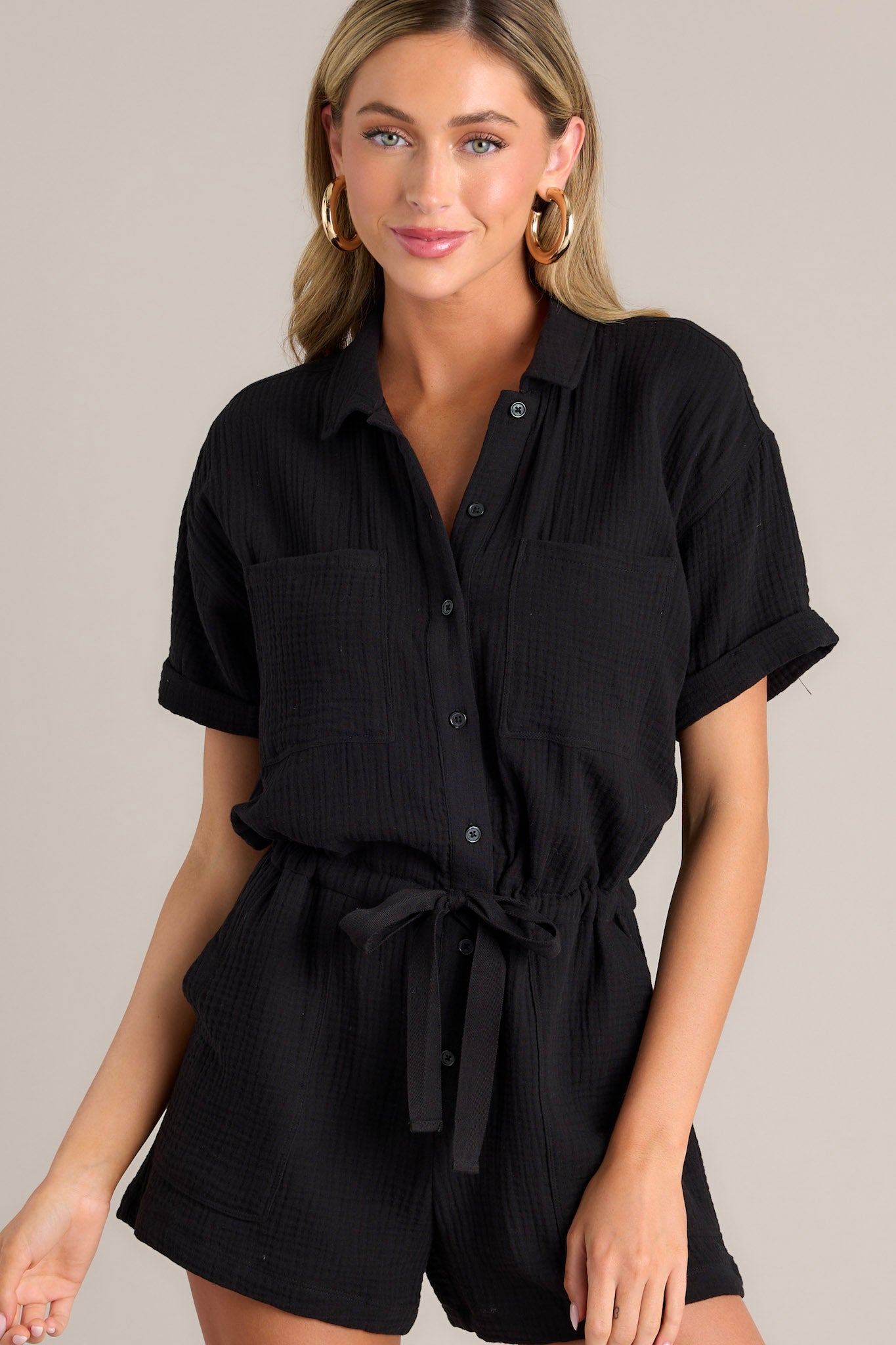 Front view of this black romper that features a collared neckline, functional breast pockets, a full button front, a self-tie drawstring waist, functional hip pockets, and cuffed short sleeves.