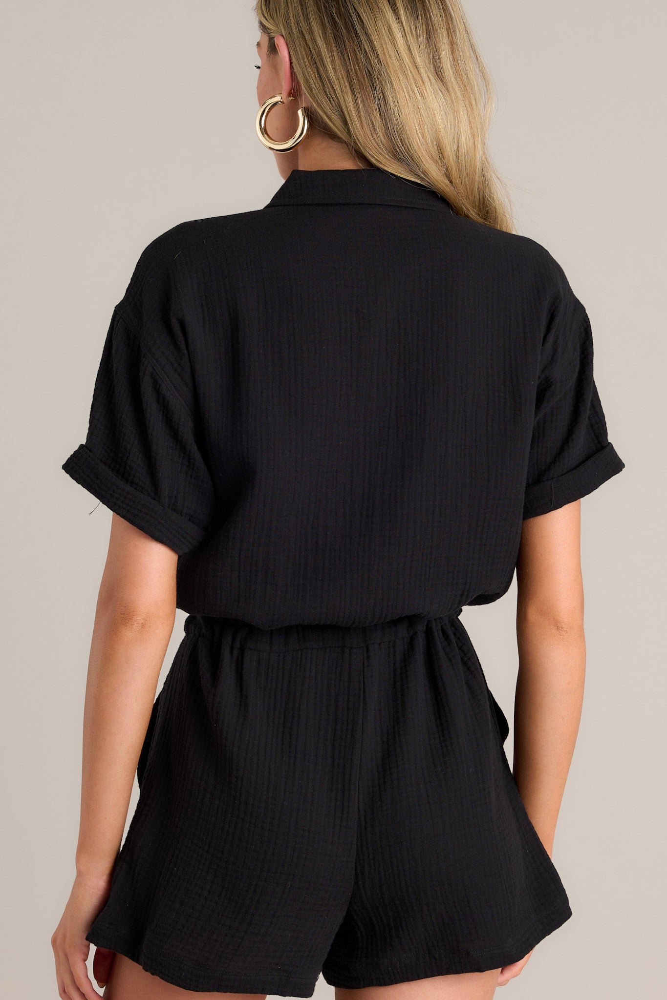 Back view of this black romper that features a collared neckline, functional breast pockets, a full button front, a self-tie drawstring waist, functional hip pockets, and cuffed short sleeves.