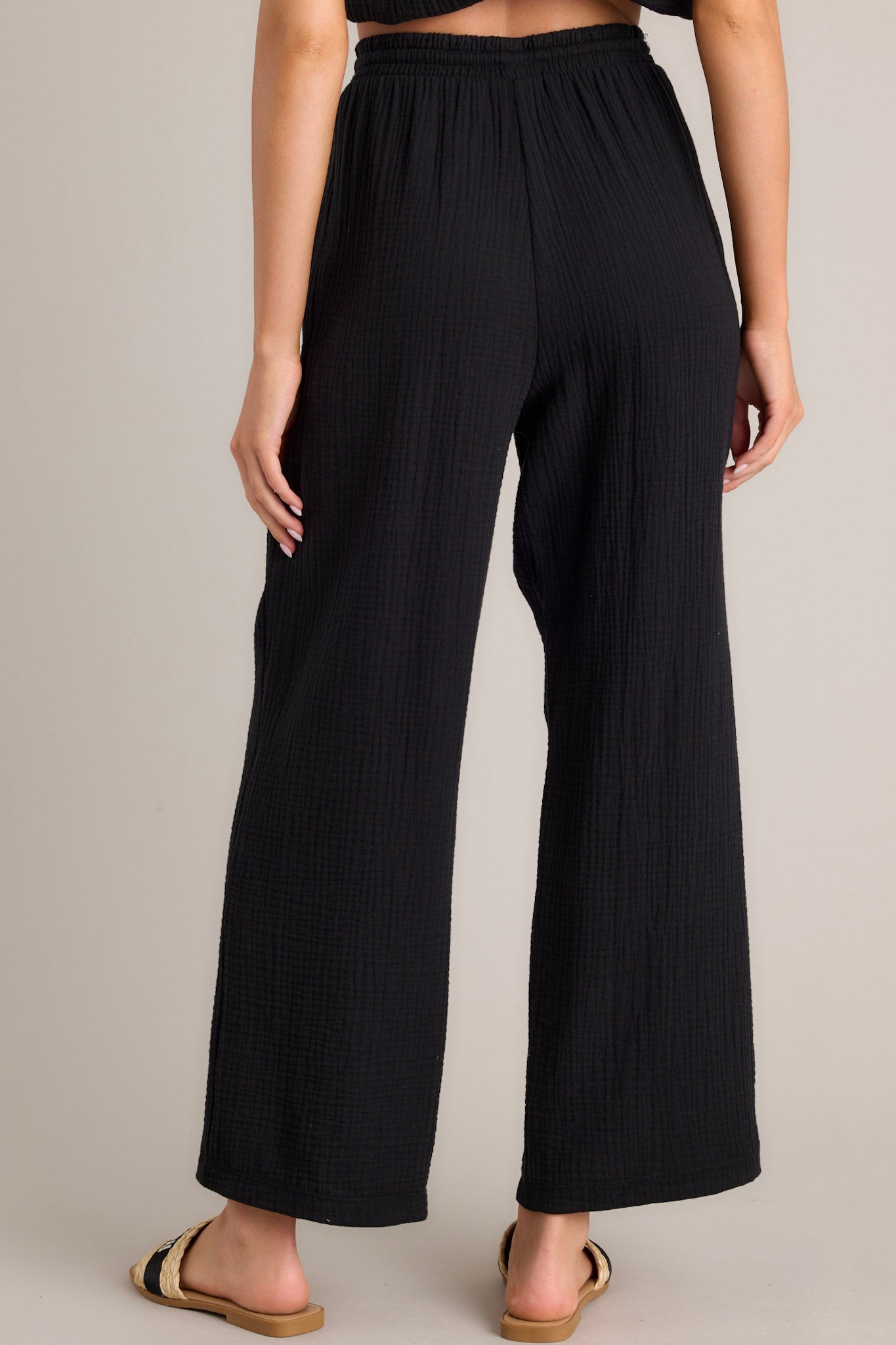 Back view of these black pants that feature a high waisted design, an elastic waistband, a self-tie drawstring, functional hip pockets, and a wide leg.