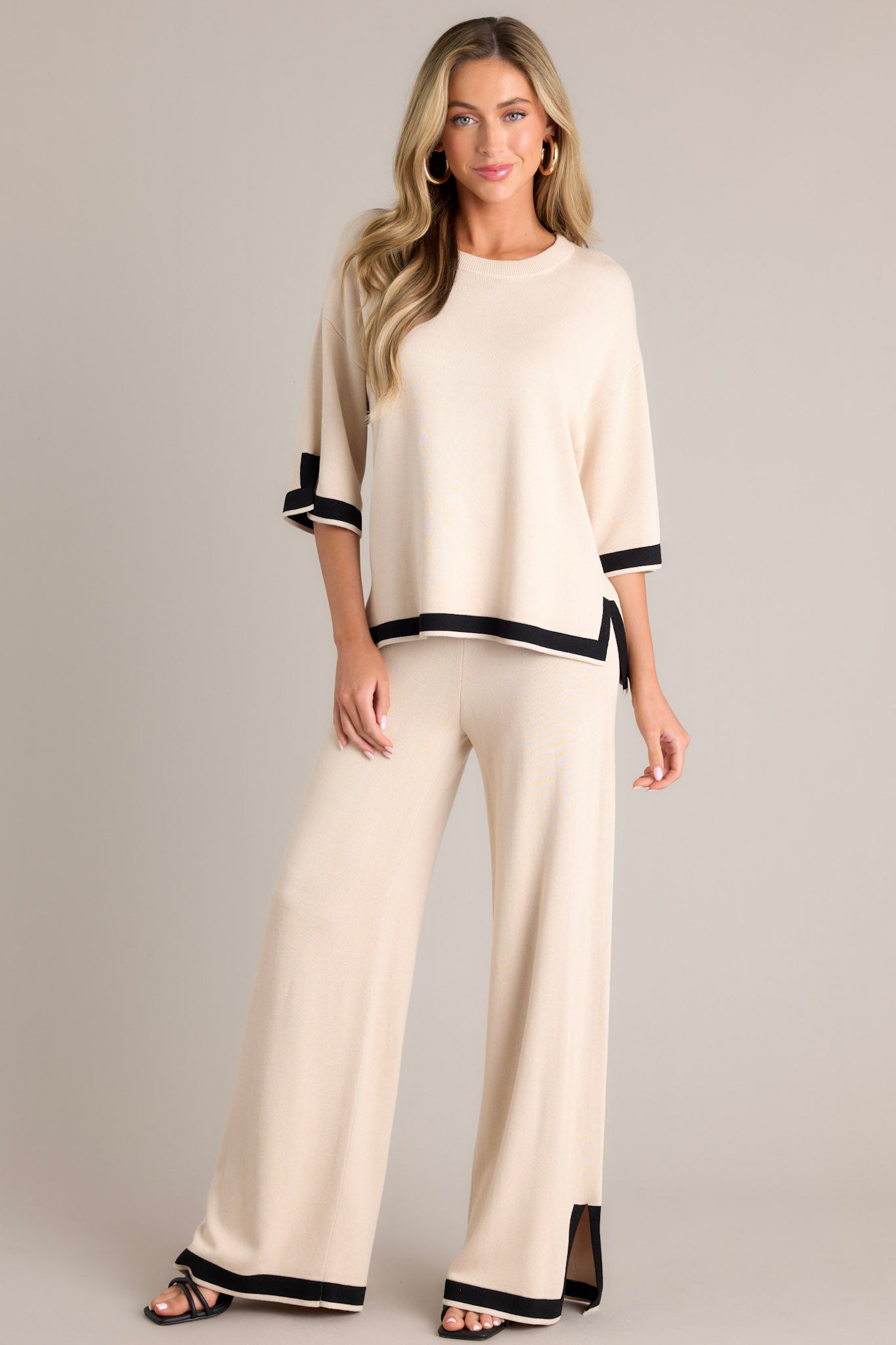 Full body view of these beige sweater pants that feature a high waisted design, an elastic waistband, a wide leg, and a contrasting split hemline.