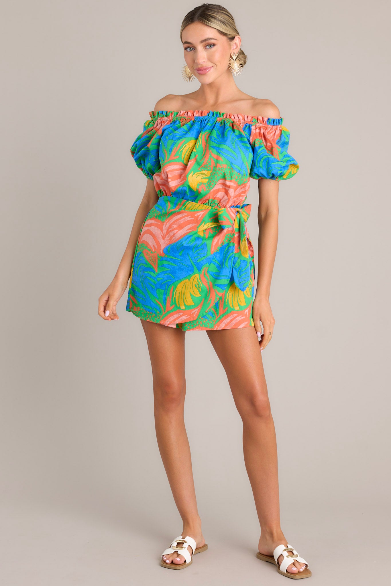 Full body view of this romper that features an elastic ruffled strapless neckline, an elastic waistband, a self-tie wrap feature, and elastic cuffed short sleeves.