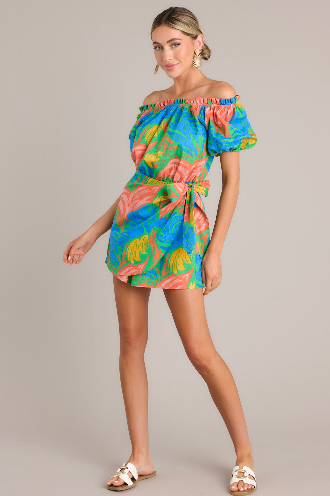 Angled view of this romper that features an elastic ruffled strapless neckline, an elastic waistband, a self-tie wrap feature, and elastic cuffed short sleeves.