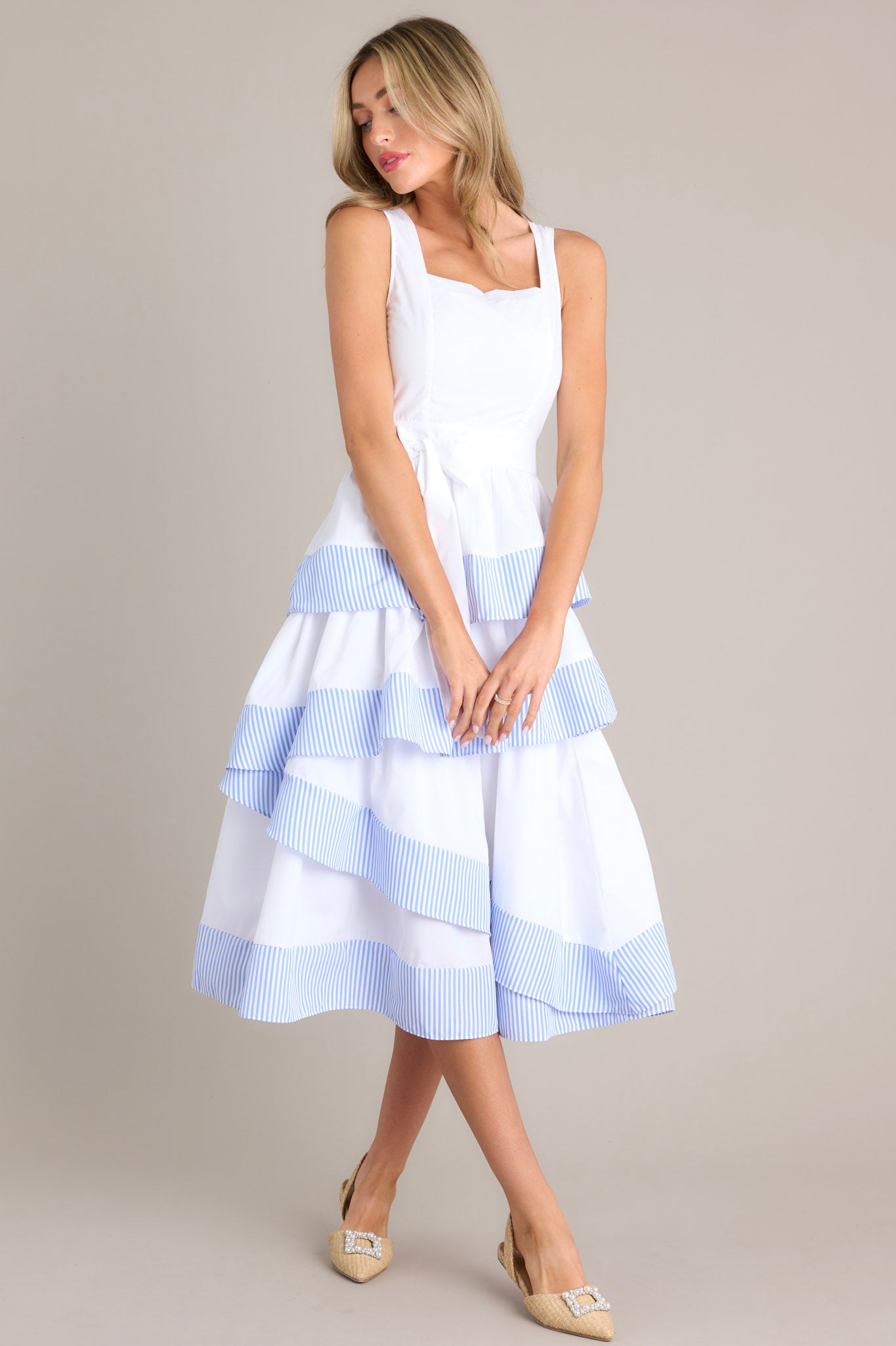 This white and blue dress features a square neckline, thick straps, a smocked back insert, a discrete zipper, a self-tie belt, belt loops, and an asymmetrical tiered design.