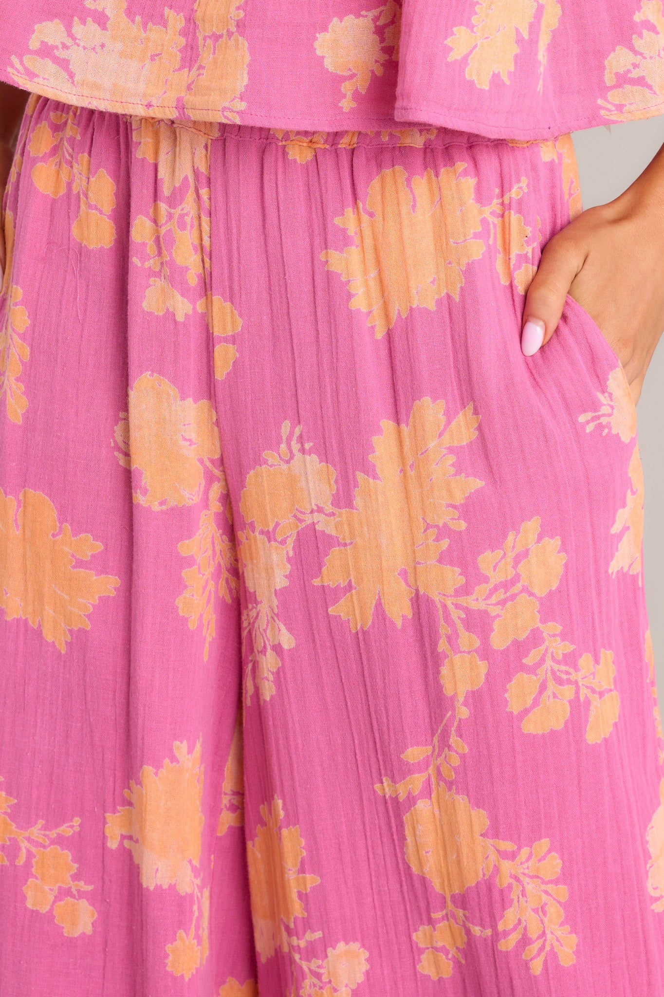 Close up view of these pink floral pants that feature a high waisted design, a thick elastic waistband, functional hip pockets, and a wide leg.