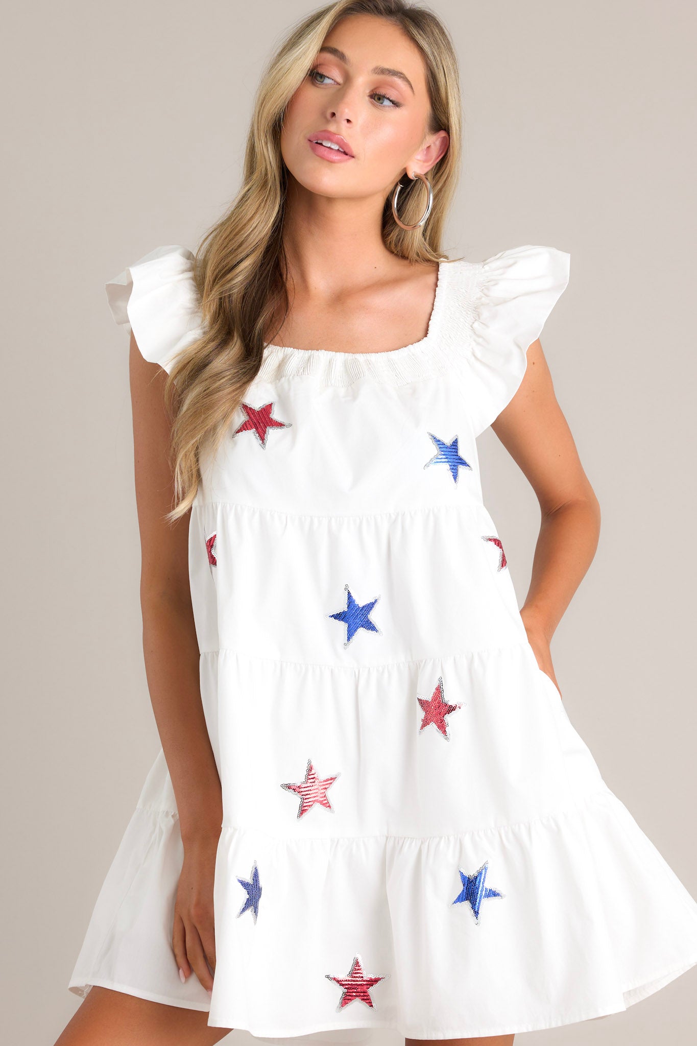 Angled front view of this white mini dress that features a smocked square neckline, multicolored sequin stars, a tiered design, and functional hip pockets.