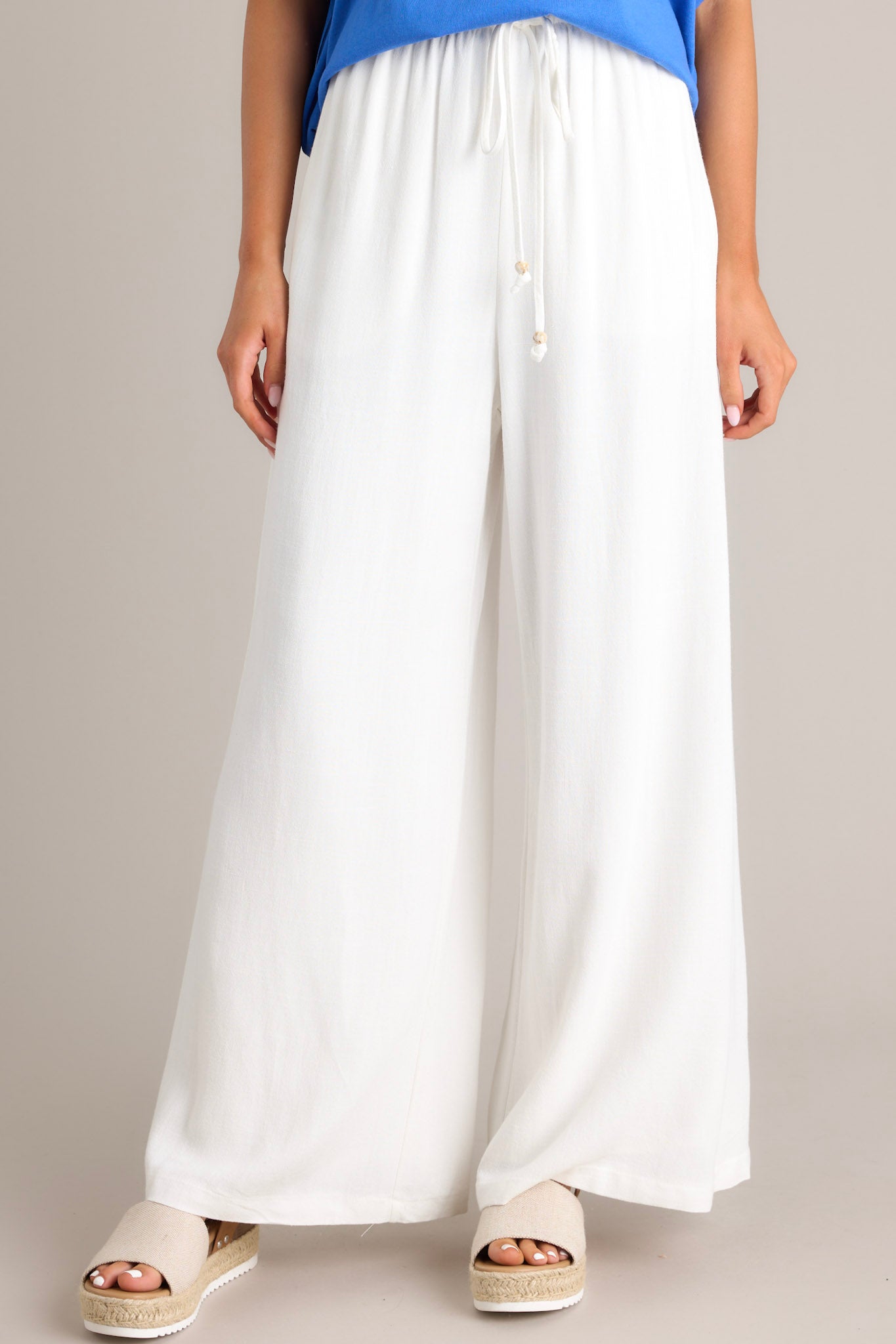 Front view of these white pants that feature a high waisted design, a thin elastic waistband, a self-tie drawstring, functional hip pockets, and a wide leg.