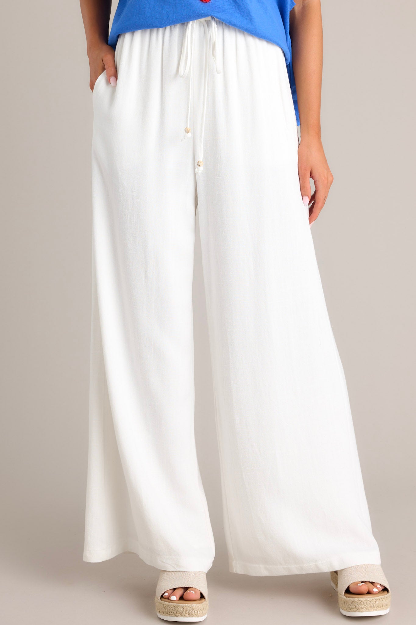 Angled front view of these white pants that feature a high waisted design, a thin elastic waistband, a self-tie drawstring, functional hip pockets, and a wide leg.