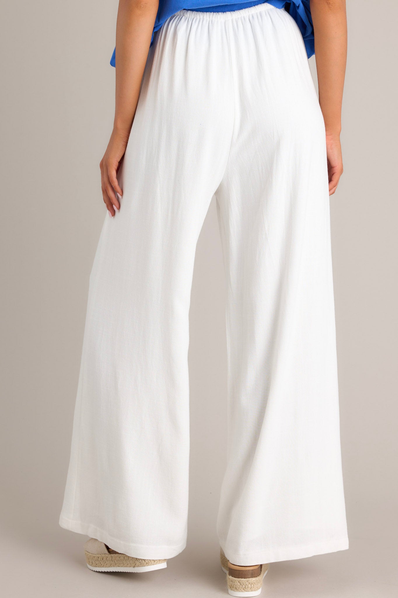 Back view of these white pants that feature a high waisted design, a thin elastic waistband, a self-tie drawstring, functional hip pockets, and a wide leg.