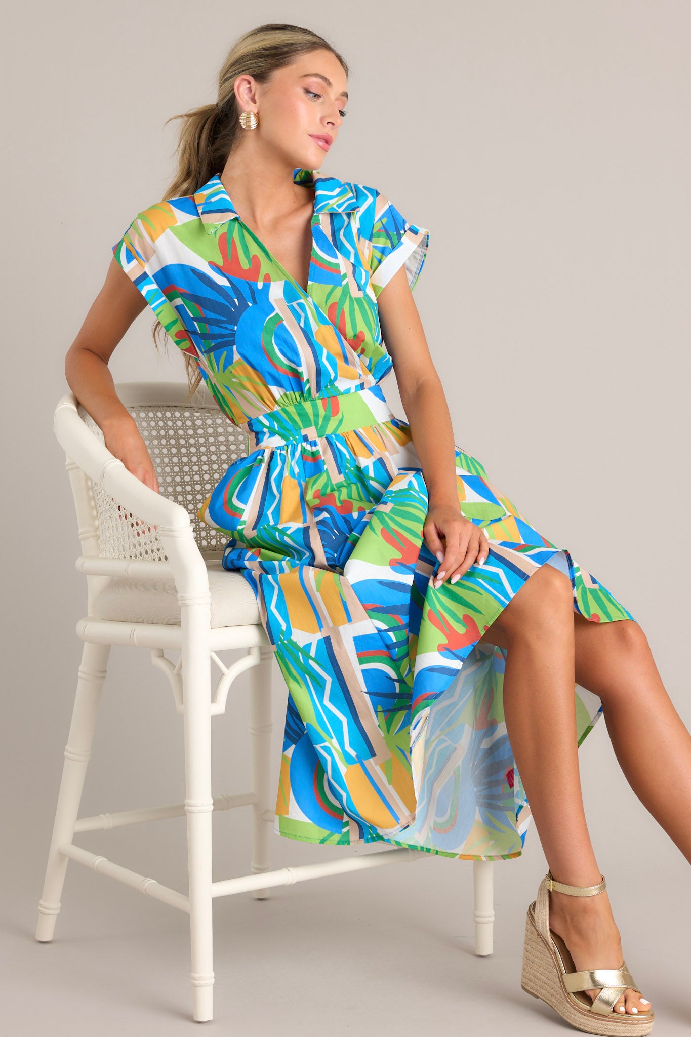 This blue midi dress features a collared v-neckline, a functional snap button, a thick waistband with a smocked insert, functional hip pockets, an abstract tropical print, and wide cuffed short sleeves.