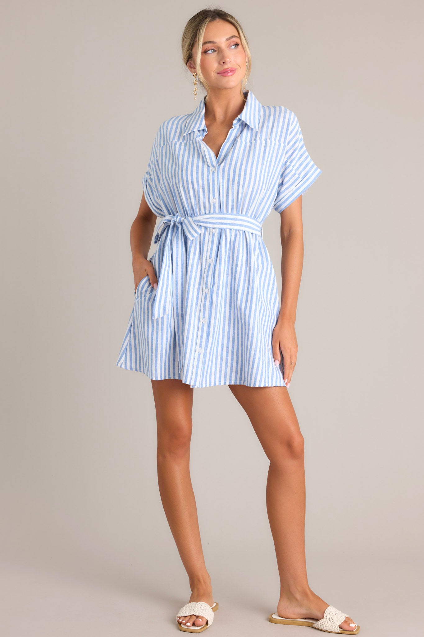 Full body view of  this dress that features a collared v-neckline, a full button front, an elastic waistband, a self-tie belt, functional pockets, and folded sleeves.