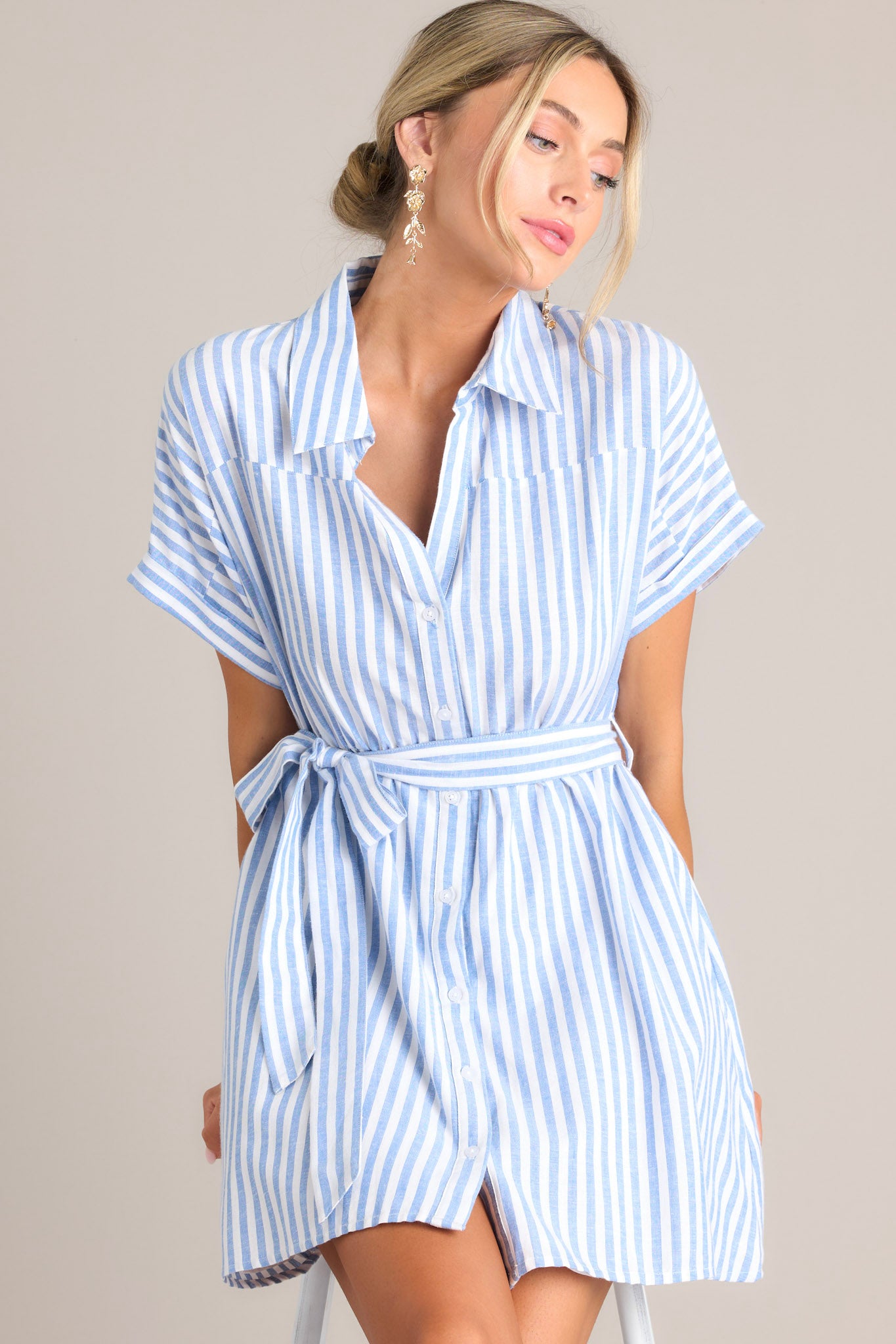 Seated front view of  this dress that features a collared v-neckline, a full button front, an elastic waistband, a self-tie belt, functional pockets, and folded sleeves.