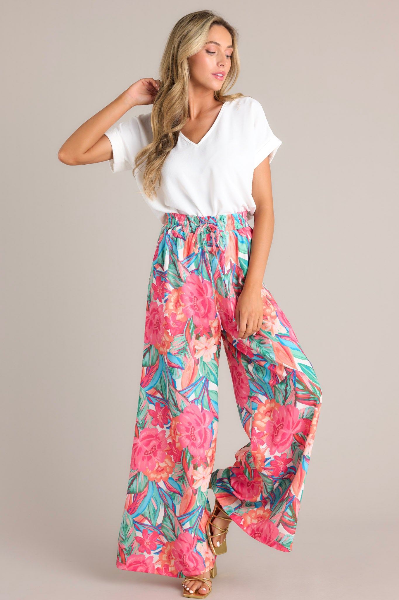 Full body view of these pink floral pants that feature a high waisted design, an elastic waistband, a self-tie drawstring, a tropical floral print, and a wide leg design.