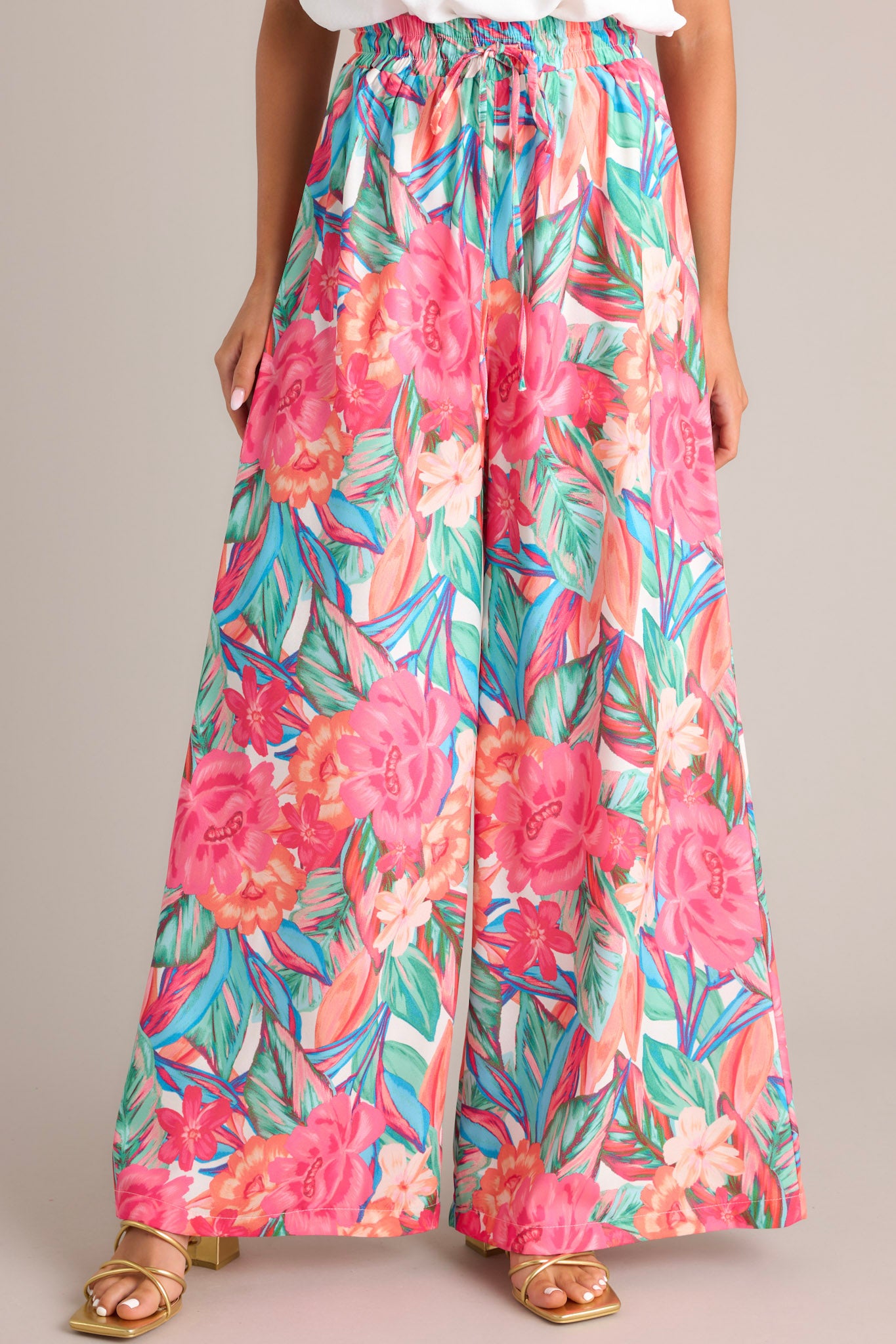 Front view of these pink floral pants that feature a high waisted design, an elastic waistband, a self-tie drawstring, a tropical floral print, and a wide leg design.