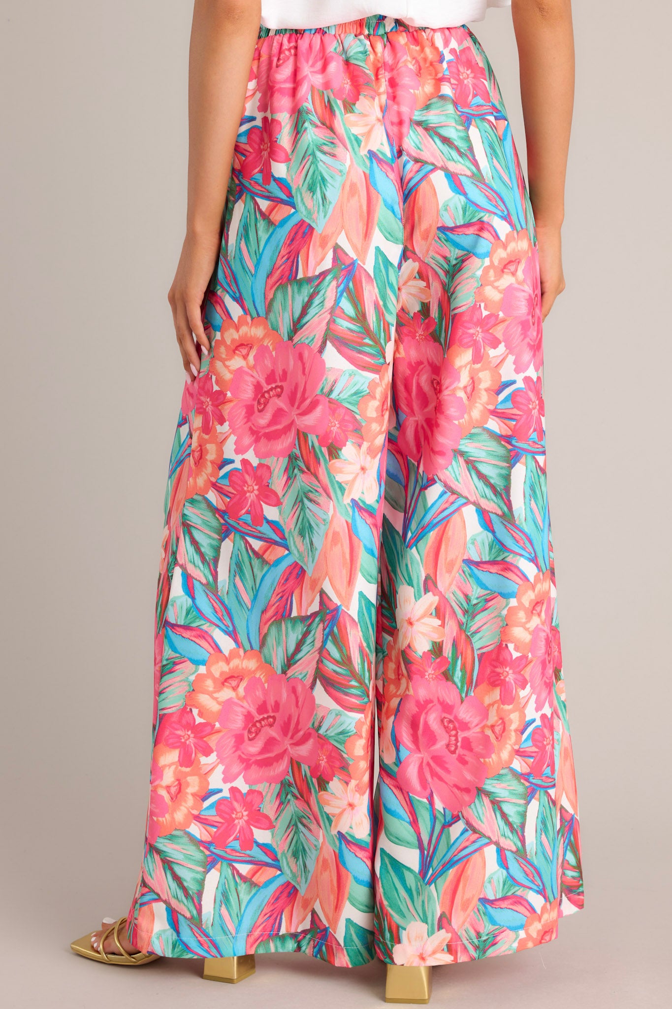 Back view of these pink floral pants that feature a high waisted design, an elastic waistband, a self-tie drawstring, a tropical floral print, and a wide leg design.