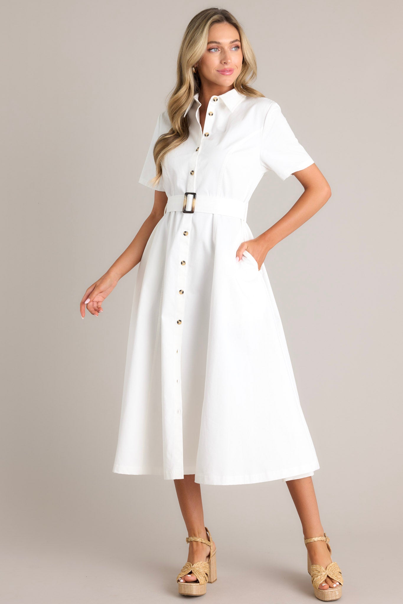 Angled full body view of this white midi dress that features a collared neckline, a full button front, a functional pull-through belt, functional hip pockets, and short sleeves.