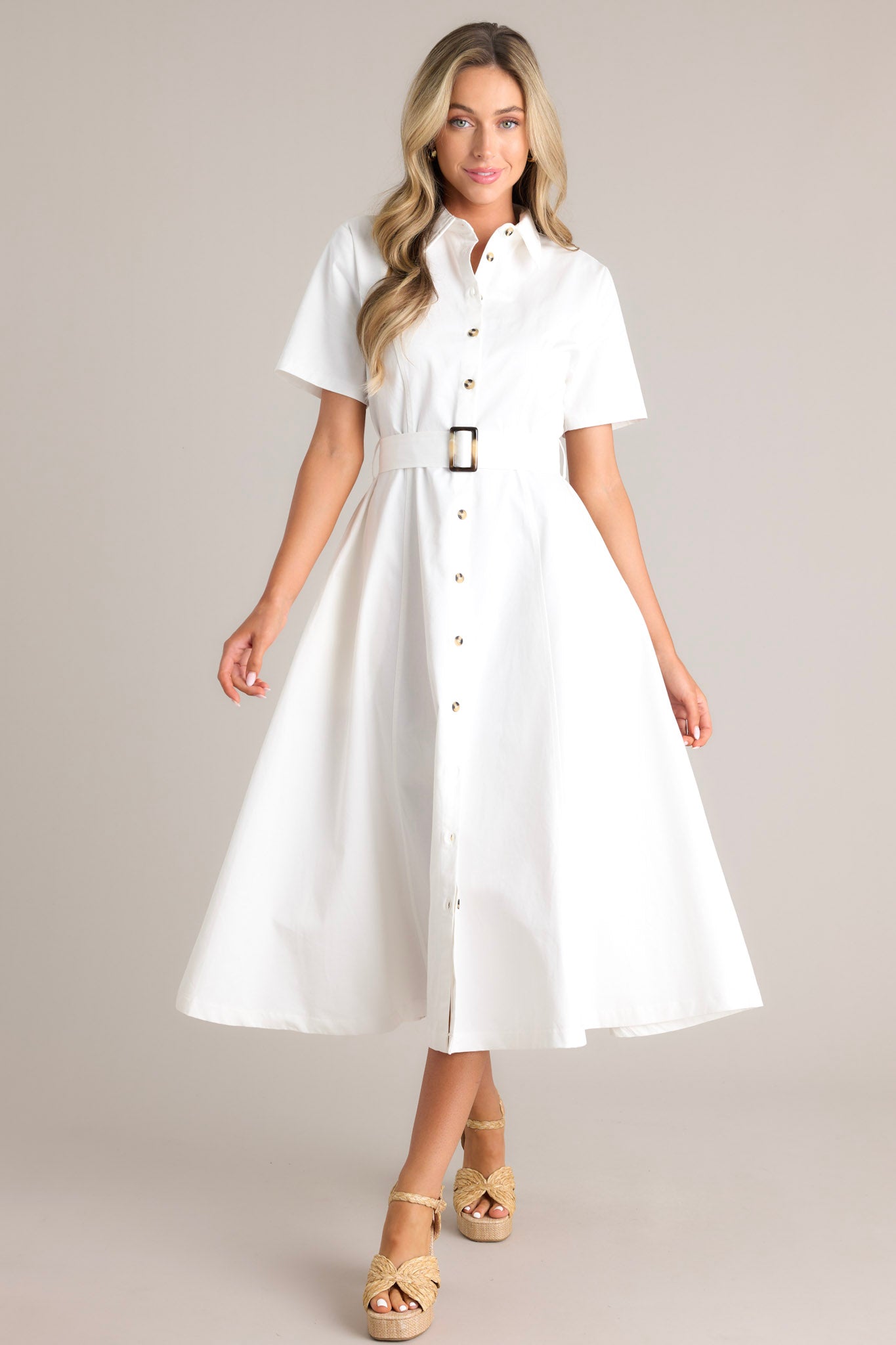 Full body view of this white midi dress that features a collared neckline, a full button front, a functional pull-through belt, functional hip pockets, and short sleeves.