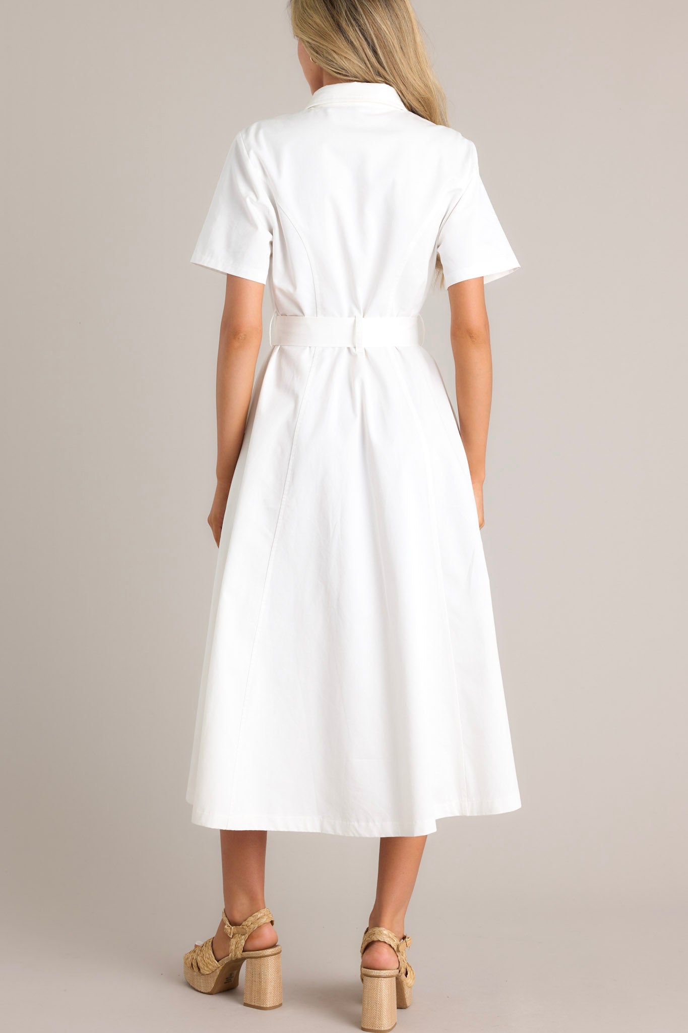 Back view of this white midi dress that features a collared neckline, a full button front, a functional pull-through belt, functional hip pockets, and short sleeves.