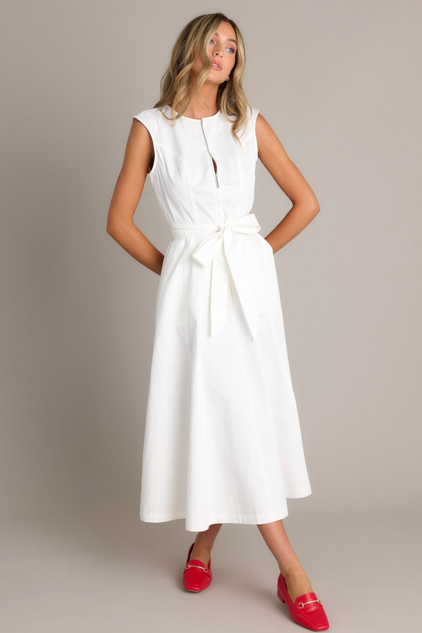 A white sleeveless midi dress with a front zipper, a fitted bodice, and a wide waist tie that forms a bow.