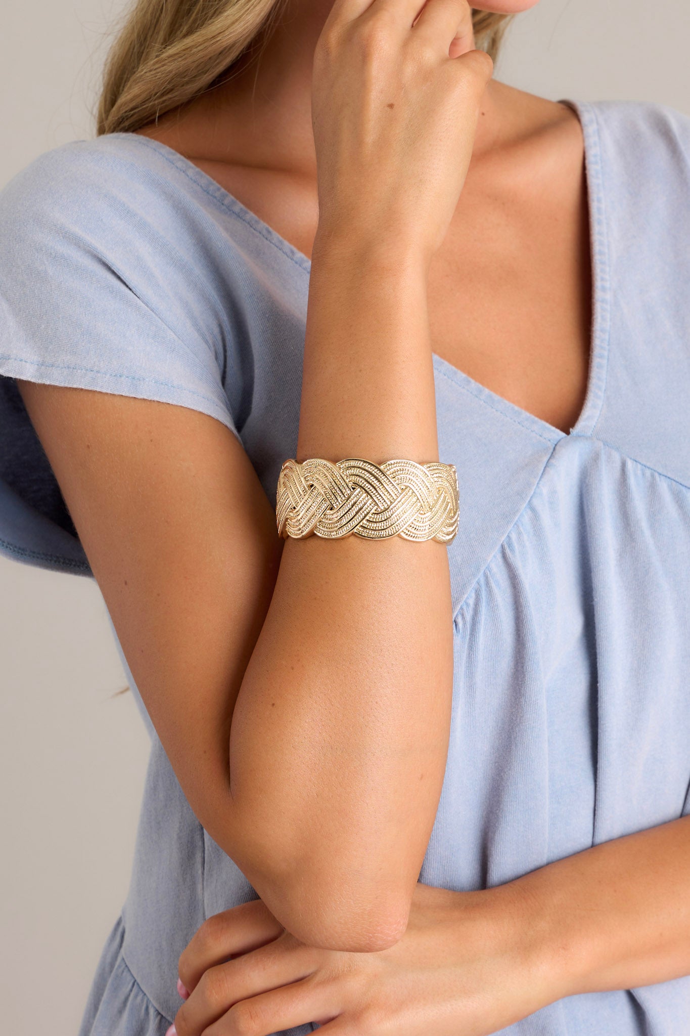 This gold cuff features a wide band, a textured braided design, and a spring hinged closure.