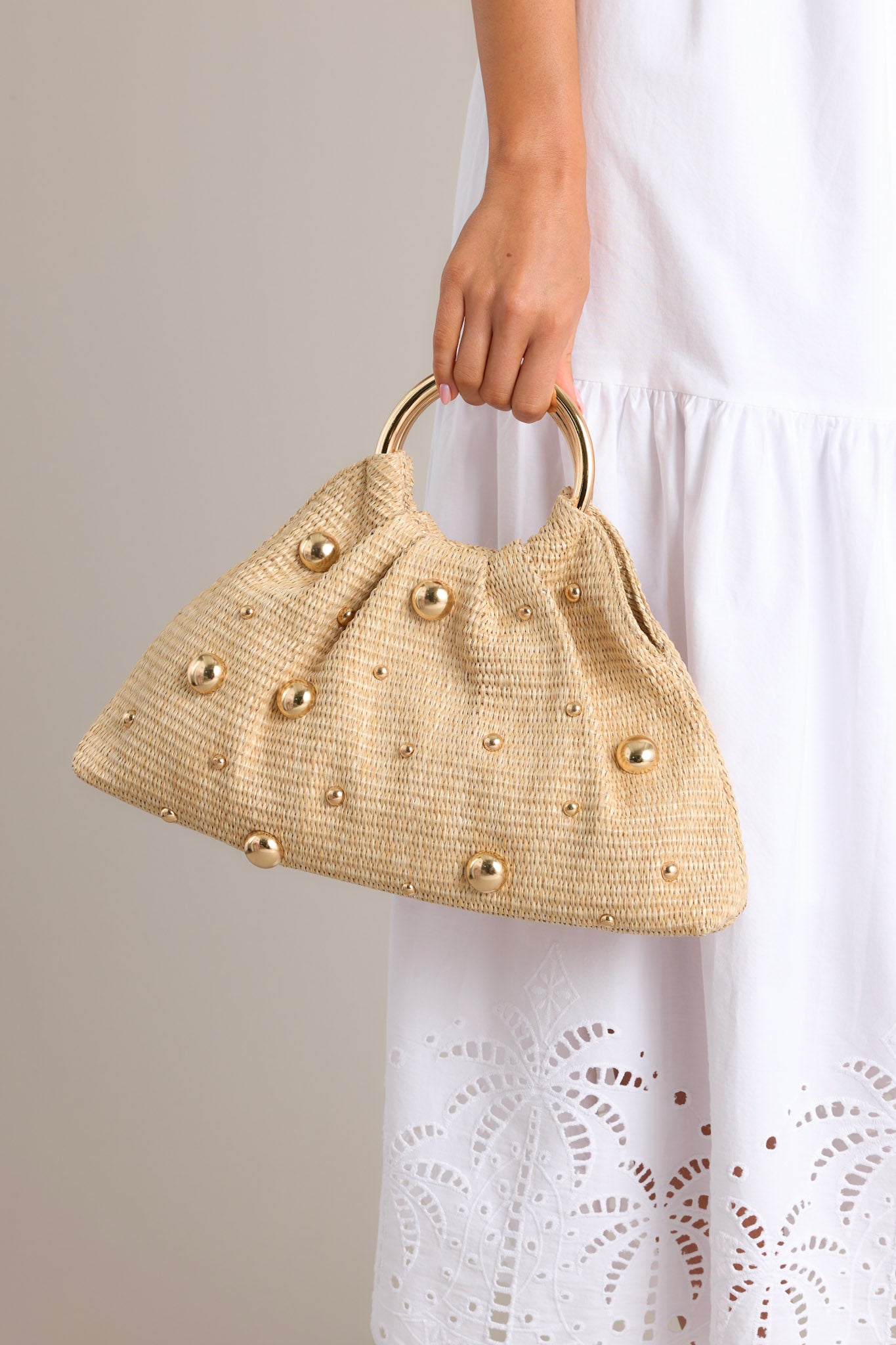 Close up of this rattan clutch that features round shiny gold handles, a magnetic closure, a spacious interior with a smaller pocket inside, and round shiny gold embellishments that vary in size.