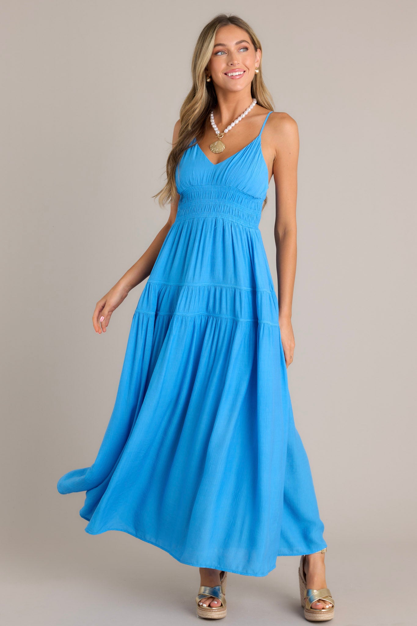 Action shot of a blue maxi dress highlighting the v-neckline, thin adjustable straps, fully smocked waist and lower back, tiers, and the flowing silhouette.