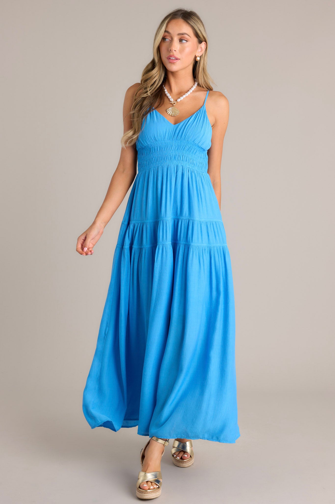 Front angled view of a blue maxi dress featuring a v-neckline, thin adjustable straps, a fully smocked waist, tiers, and a flowing silhouette