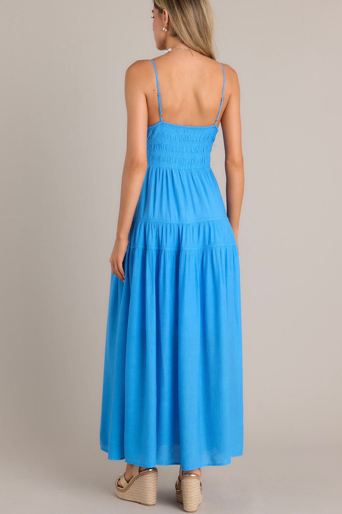 Back view of a blue maxi dress showcasing an open back, thin adjustable straps, and a fully smocked lower back.