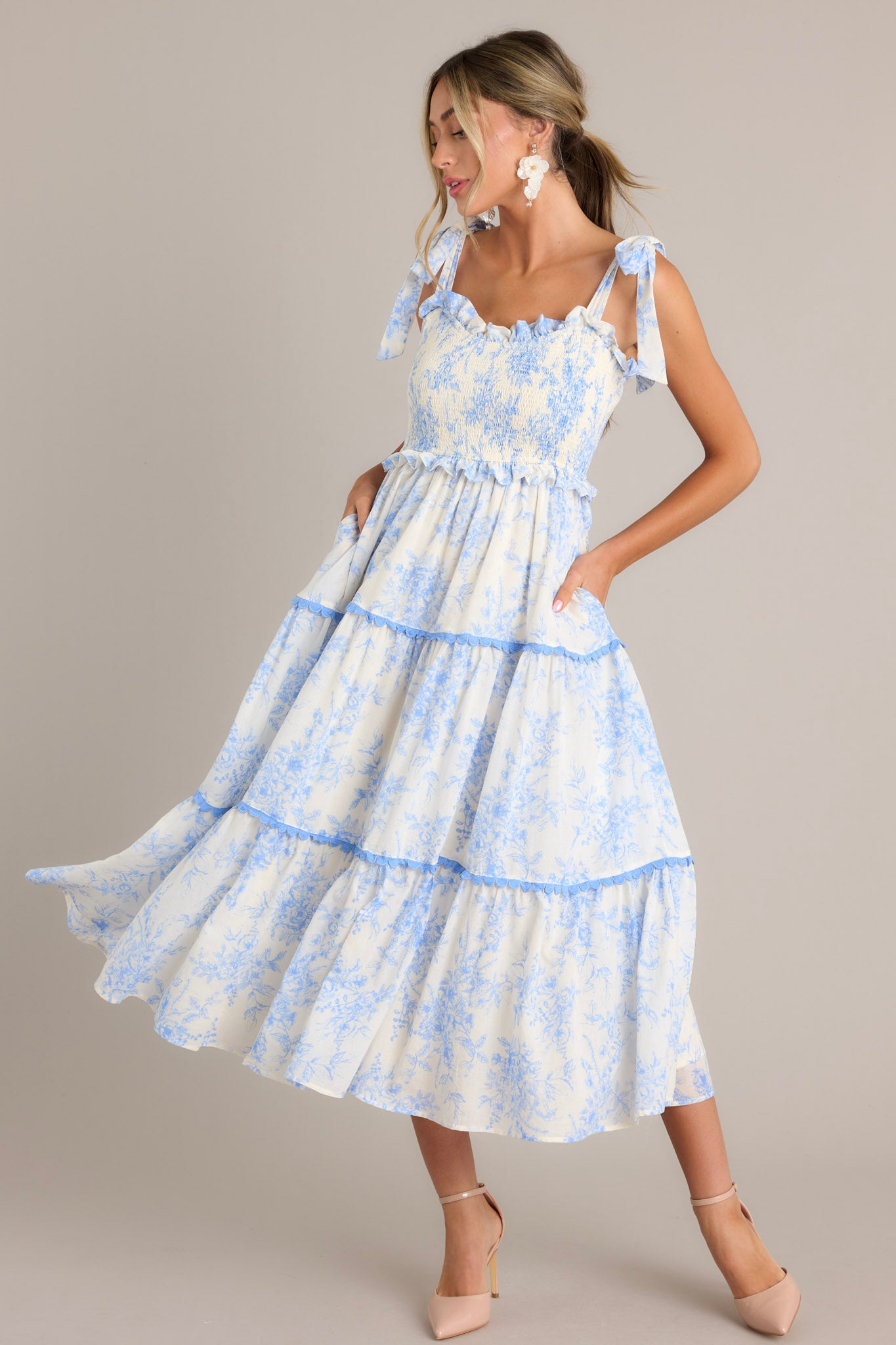 Angled full body view of this light blue midi dress that features a ruffled sweetheart neckline, self-tie straps, a fully smocked bodice, functional hip pockets, and scalloped tiers.