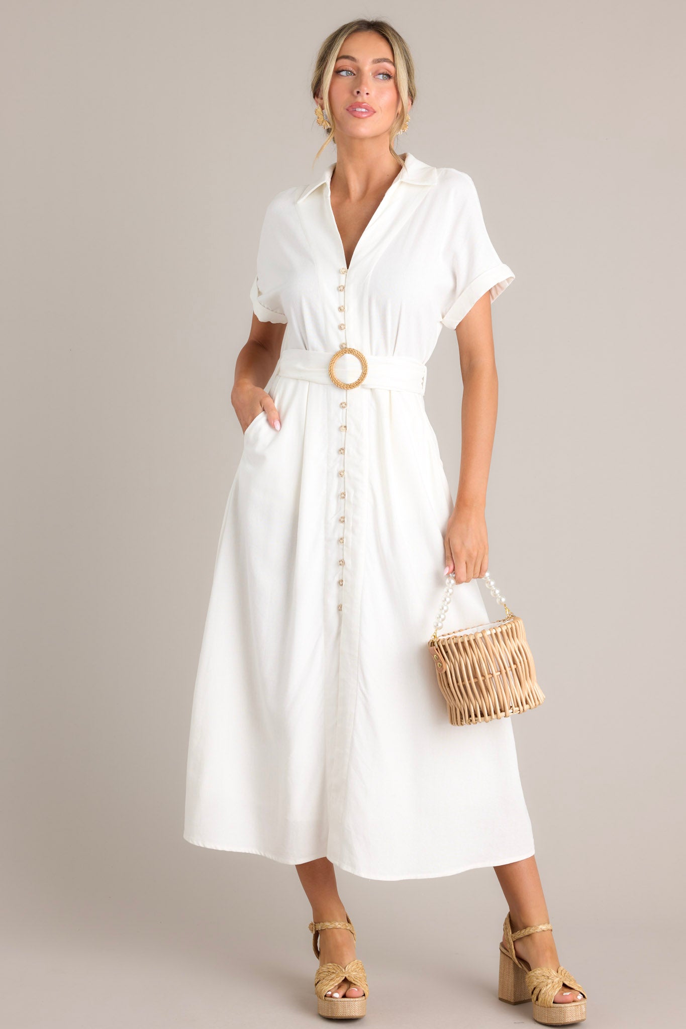 This white maxi dress features a collared v-neckline, a functional button front, a pull-through belt, functional hip pockets, a front slit, and cuffed short sleeves.