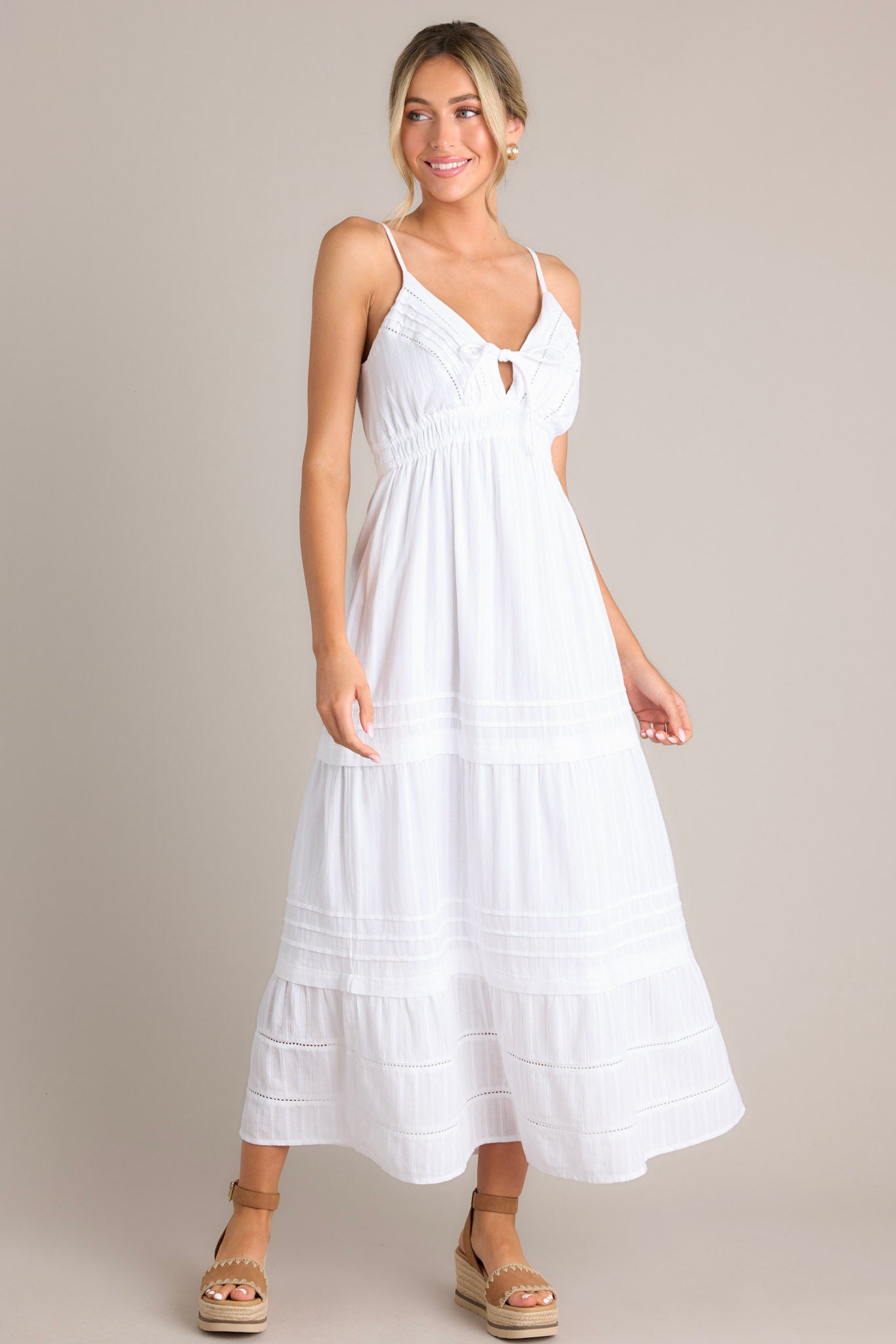 Angled full body view of this white midi dress that features a v-neckline, thin adjustable straps, eyelet bust detailing, a self-tie bust feature, an elastic waistline, and a tiered design.