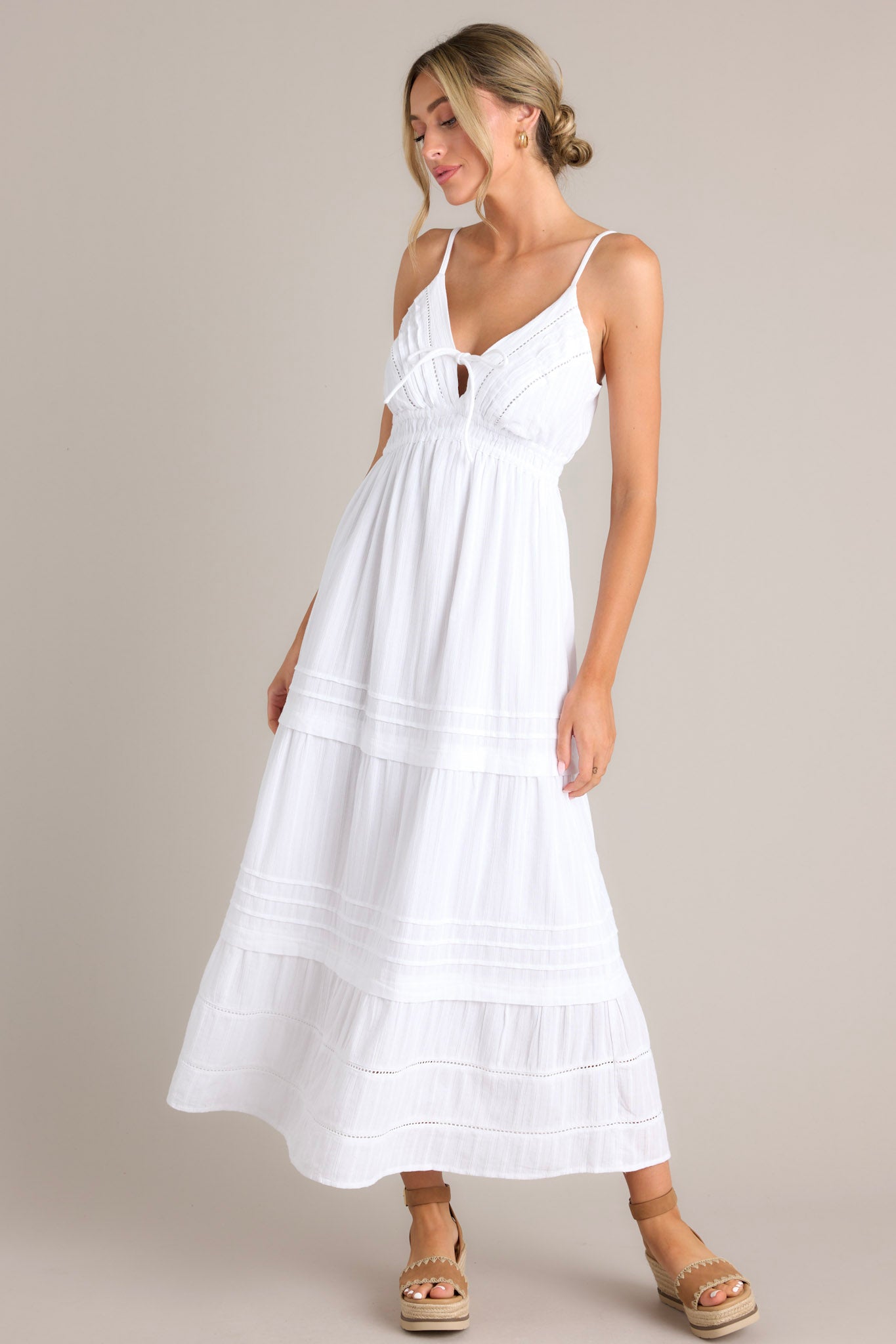 Full body view of this white midi dress that features a v-neckline, thin adjustable straps, eyelet bust detailing, a self-tie bust feature, an elastic waistline, and a tiered design.