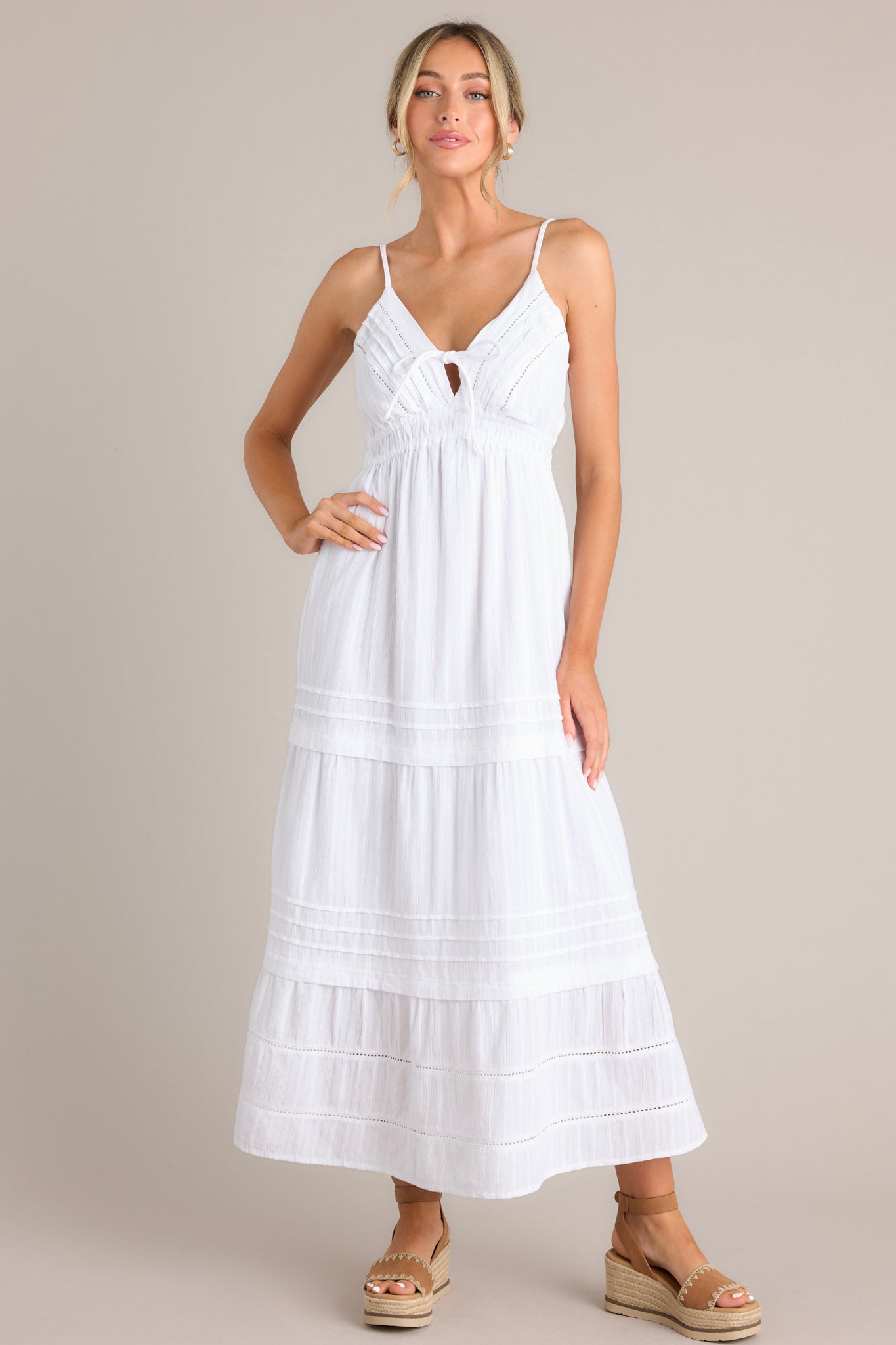 Full body front view of this white midi dress that features a v-neckline, thin adjustable straps, eyelet bust detailing, a self-tie bust feature, an elastic waistline, and a tiered design.