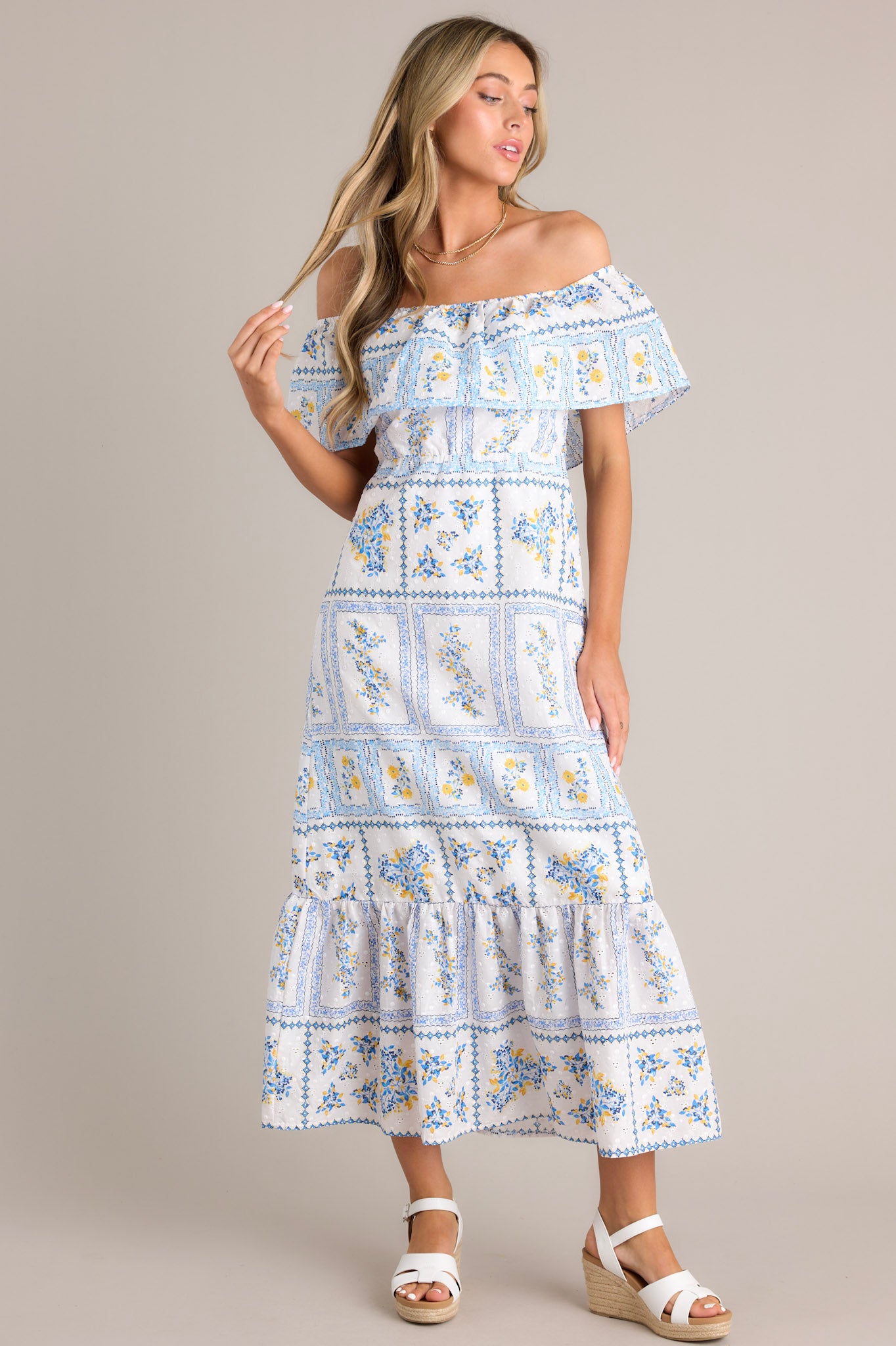 Full front view of this white & blue maxi dress featuring an elastic off-the-shoulder neckline, a frilled overlay that forms a false sleeve, an elastic stretch waist, an embroidered floral design, and a lined and tiered skirt.