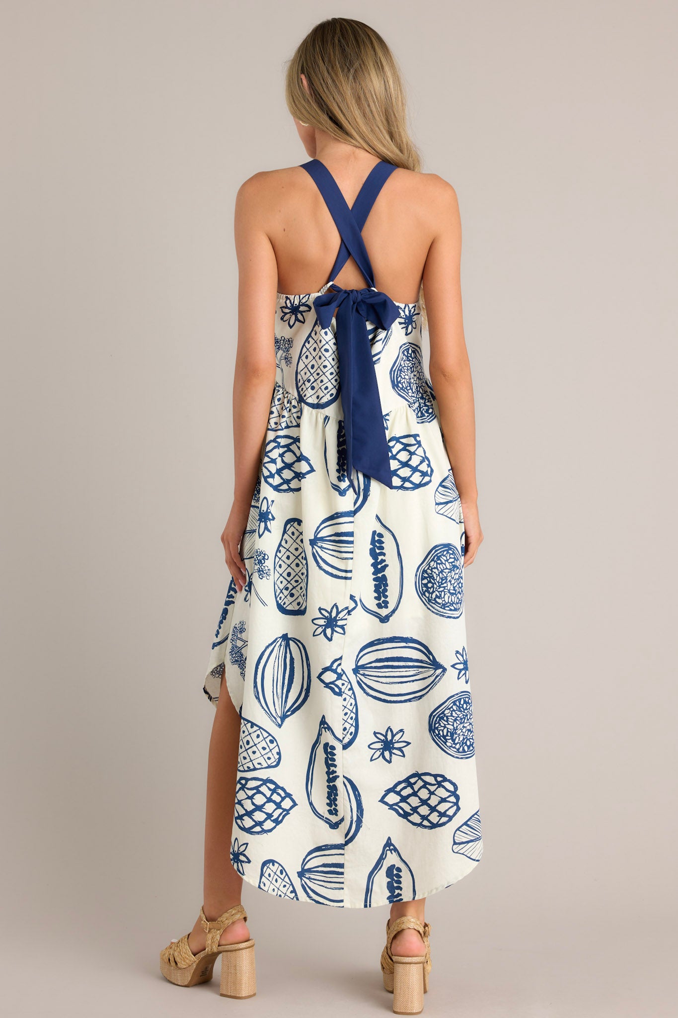 Back view of this navy midi dress that features a square neckline, self-tie straps that cross in the back, an open back, an elastic insert in the back of the waist, functional hip pockets, a unique print, and a scoop hemline.