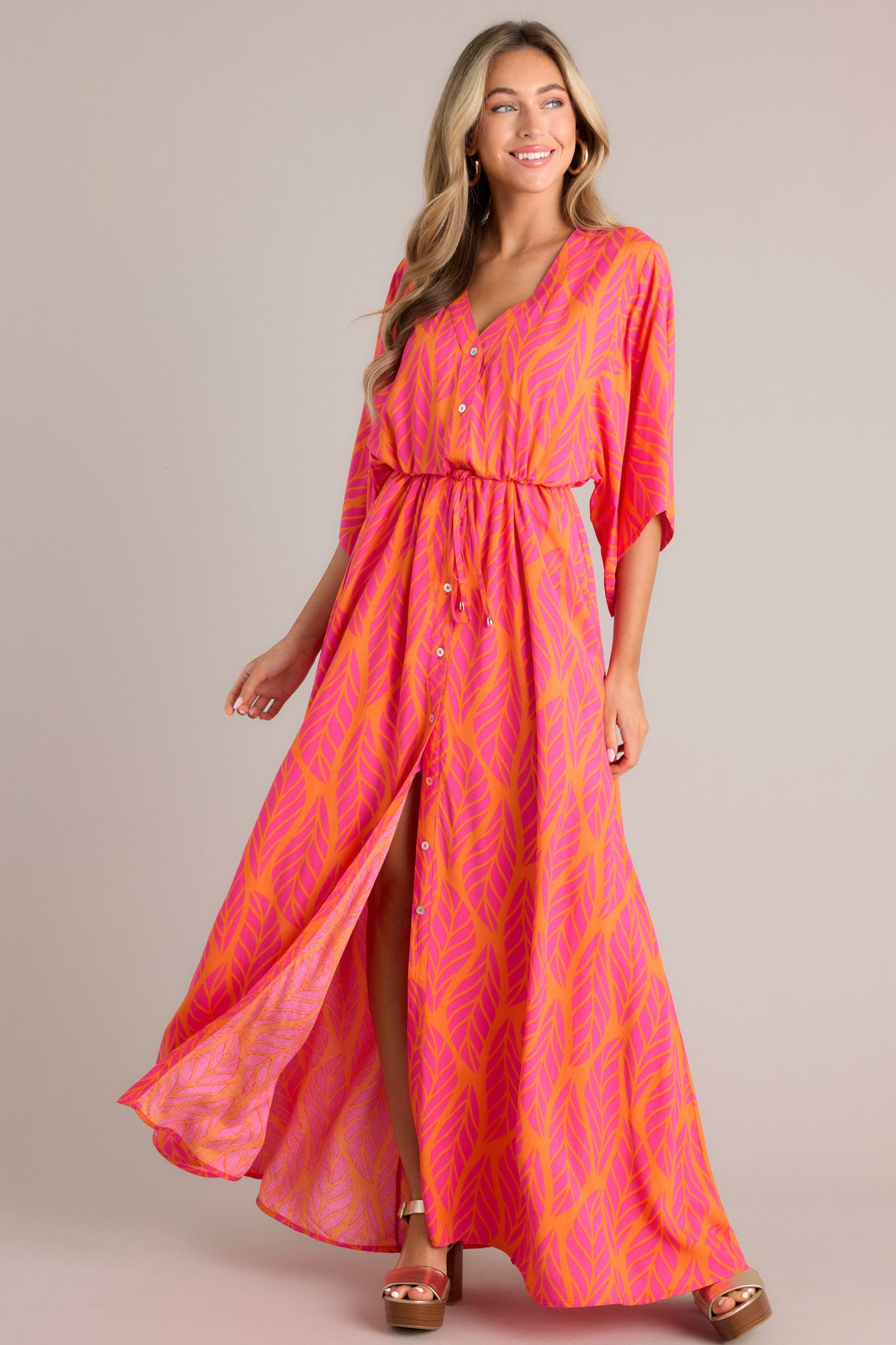 Angled full body view of this hot pink maxi dress that features a v-neckline, a functional button front, a self-tie drawstring waist, a flowing silhouette, and flowing half sleeves.